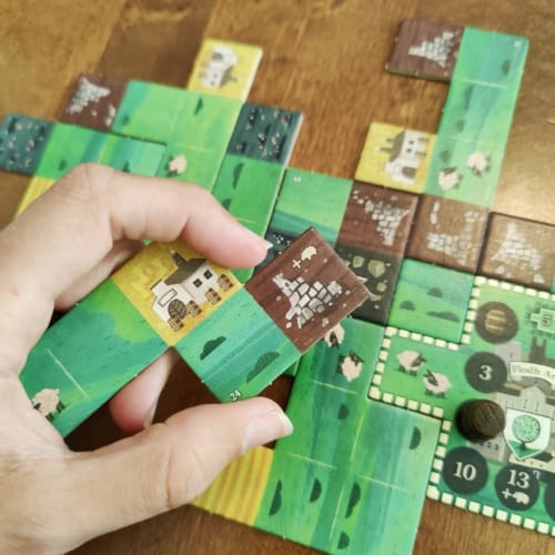 Käsi pitelee laattaa, jossa on raunio ja tislaamo laatoista rakentuvan pelialueen yläpuolella.