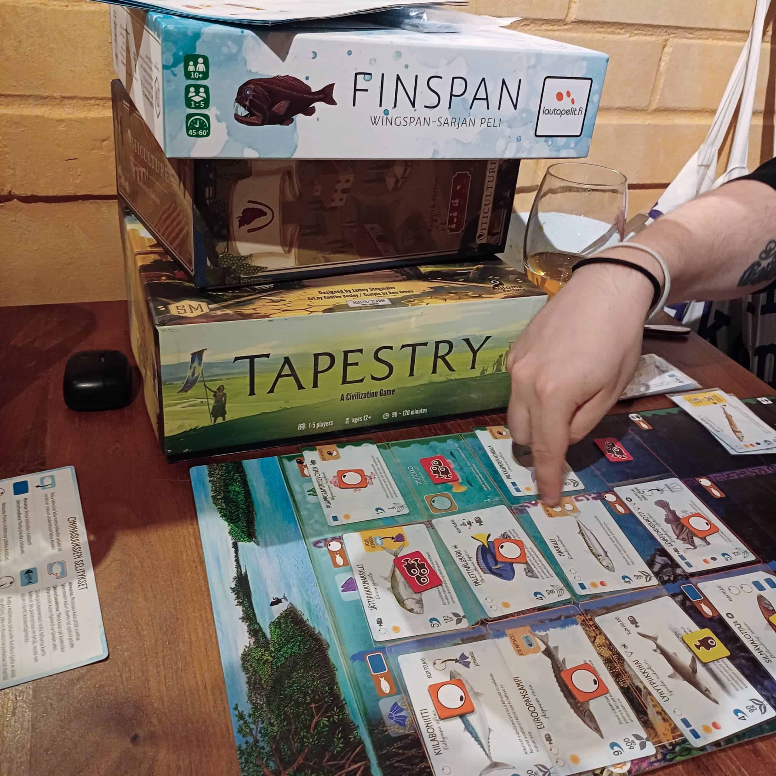Pöydällä pinossa Tapestryn, Viticulturen ja Finspanin laatikot. Etualla Finspanin pelilauta, jossa on kalakortteja, ja pelaajan käsi osoittamassa Makrilli-korttia.