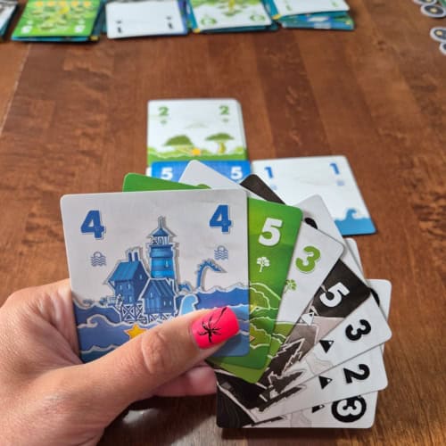Paper World -pelin kortteja kädessä. Korteissa on kuvattu paperimaisemia: esimerkiksi vettä, josta nousee rakennuksia ja merihirviö. Kortteja pitelevässä kädessä on peukalossa fuksianvärinen kynsi, jossa on musta palmukuvio.