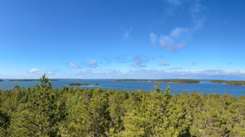 Saaristomaisemaa näkötornista: etualalla on mäntyjen latvoja, taaempana näkyy merta ja metsäisiä pikkusaaria. Taivaalla on vähän vaaleita pilviä.