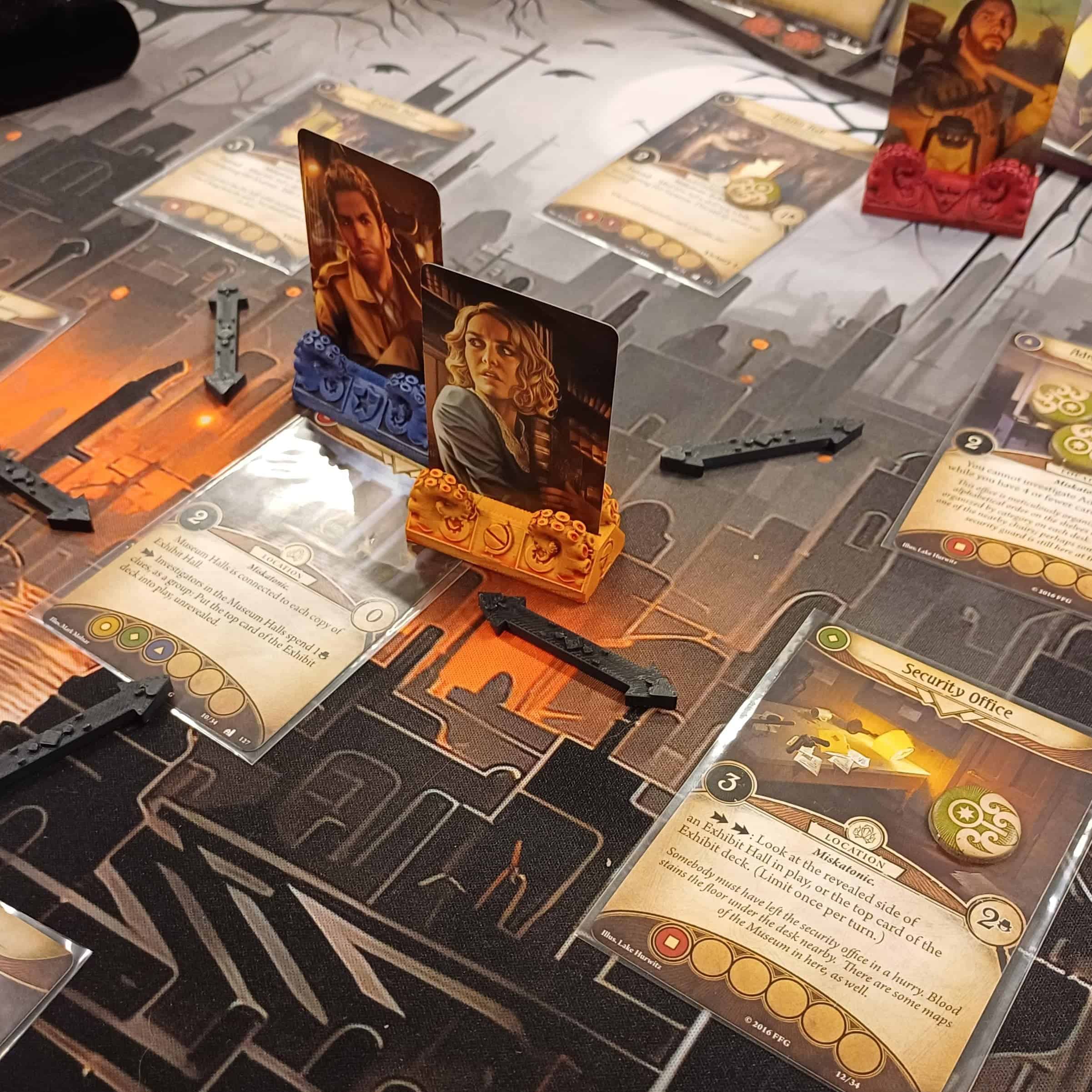 Lähikuva Arkham Horror -pelistä. Pelialustalle on pelattu paikkoja kuvaavia kortteja (museo, turvatoimisto ja muita), joiden välillä on yhteyksiä kuvaavia 3d-tulostettuja nuolia. Pelihahmoja edustavat 3d-tulostetut lonkerokuvioiset telineet, joissa on hahmojen kuvat pystyssä.