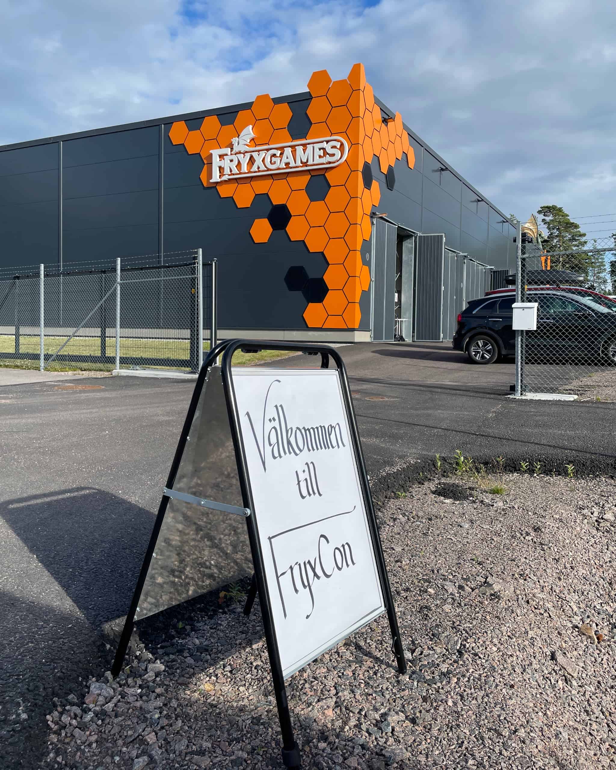 Taustalla Fryxgamesin päämajarakennus (musta teollisuushalli, jonka kulmassa oranssia heksalaattakuviointia), etualalla A-kyltti, jossa teksti "Välkommen till FryxCon".