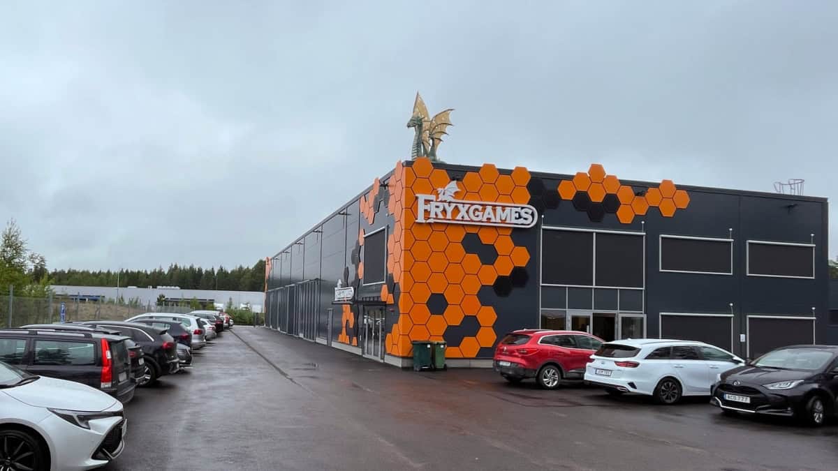 Fryxgamesin päämaja: musta teollisuusrakennus, jonka kulma on koristeltu oransseilla heksakuvioilla ja jonka katolla on lohikäärme. Pihan autopaikat ovat täynnä autoja.