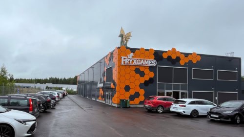 Fryxgamesin päämaja: musta teollisuusrakennus, jonka kulma on koristeltu oransseilla heksakuvioilla ja jonka katolla on lohikäärme. Pihan autopaikat ovat täynnä autoja.