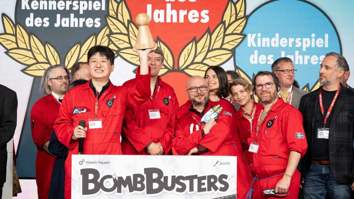 Hisashi Hayashi Spiel des Jahres -palkinnon lavalla, toisessa kädessään mikrofoni ja toisessa iso puinen pelinappula eli SdJ:n Pöppel-palkinto. Hayashin ympärillä parveilee paljon muita pelin tekemiseen liittyneitä henkilöitä, joilla kaikilla on Hayashin tapaan punaiset Bomb Busters -haalarit yllään.