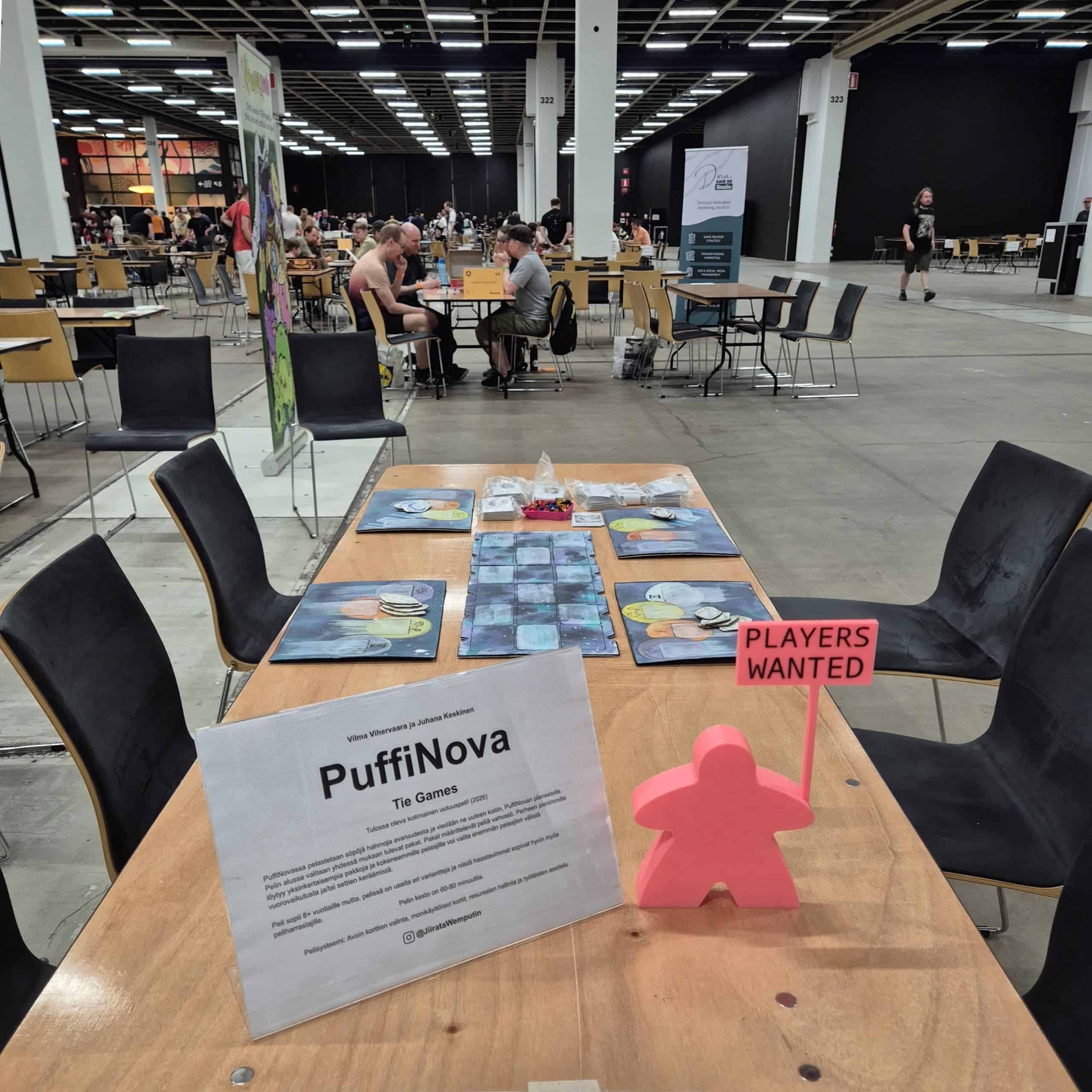 PuffiNova-pelipöytä Ropeconissa. Pöydällä on taaempana peli ja etualalla esittelykyltti ja pinkki meeplepatsas, jolla on "Players wanted"-kyltti.