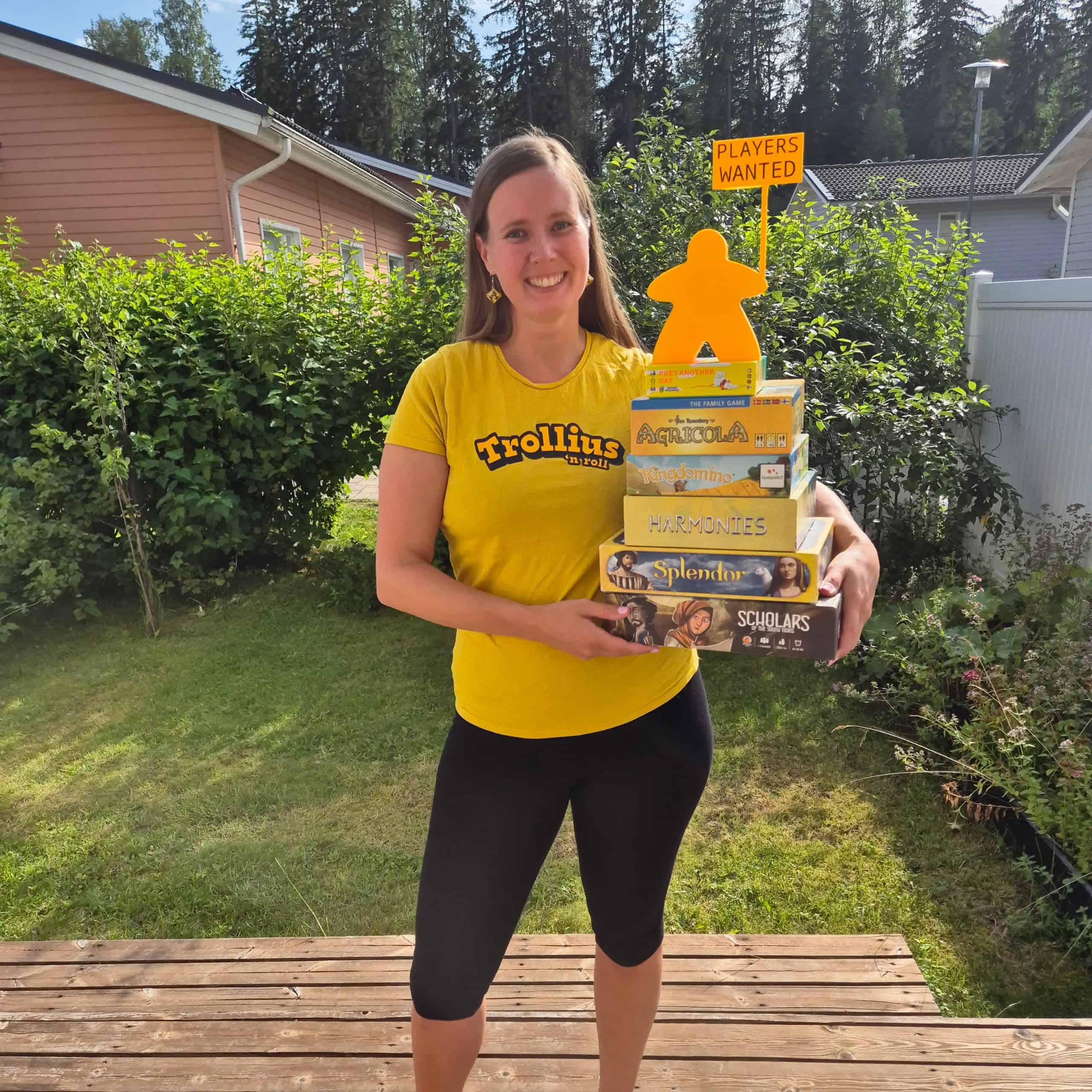 Vilma keltaisessa Trollius-paidassa, sylissään pino keltaisia pelejä: Scholars of the South Tigris, Splendor, Harmonies, Kingdomino, Agricola: The Family Game, Prey Another Day. Pinon päällä on keltainen "players wanted"-meeplepatsas.
