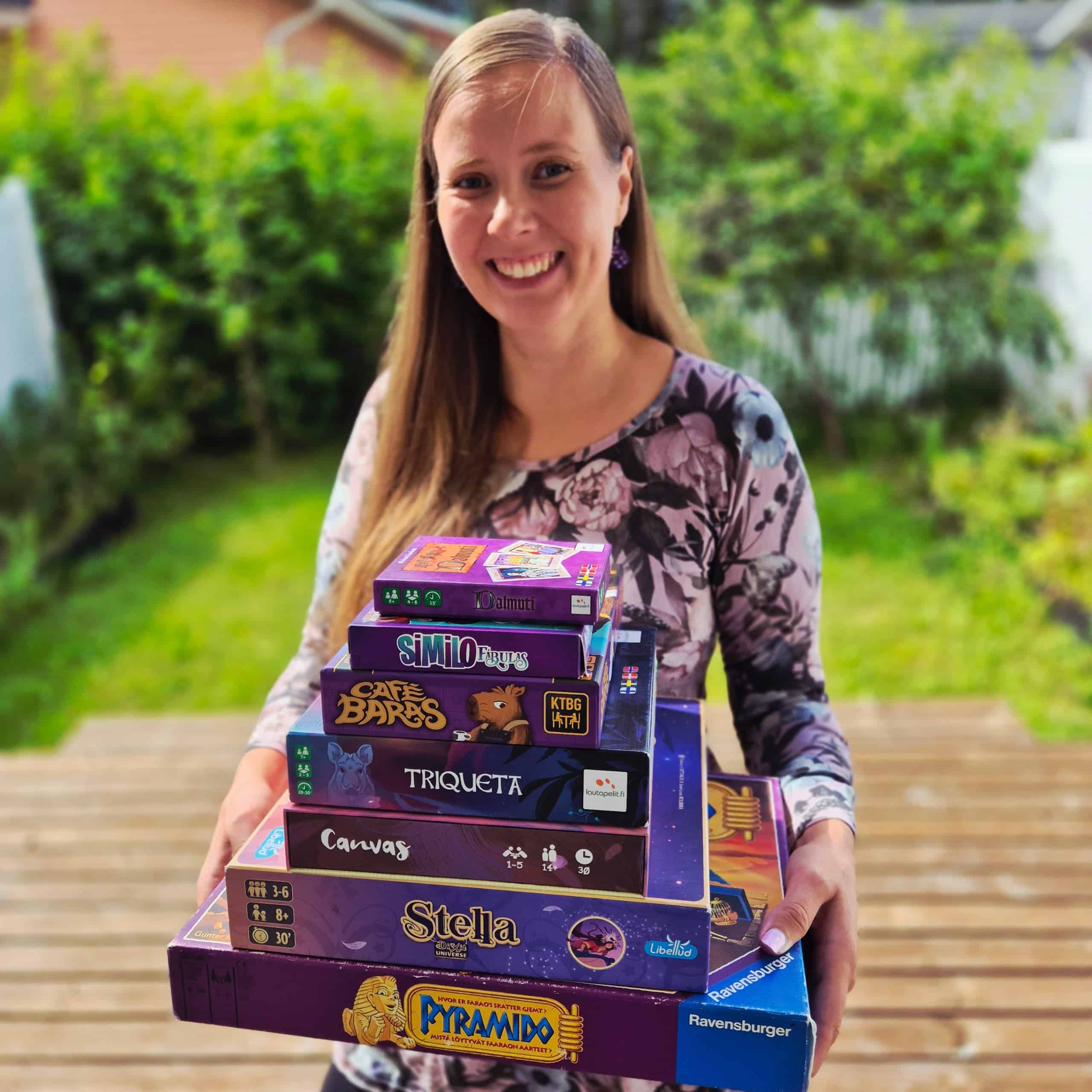 Vilma ulkona terassilla violetin sävyisissä vaatteissa ja sylissään pino violetteja pelilaatikoita: Pyramido, Stella, Canvas, Triqueta, Café Baras, Similo Fabulas ja Dalmuti.