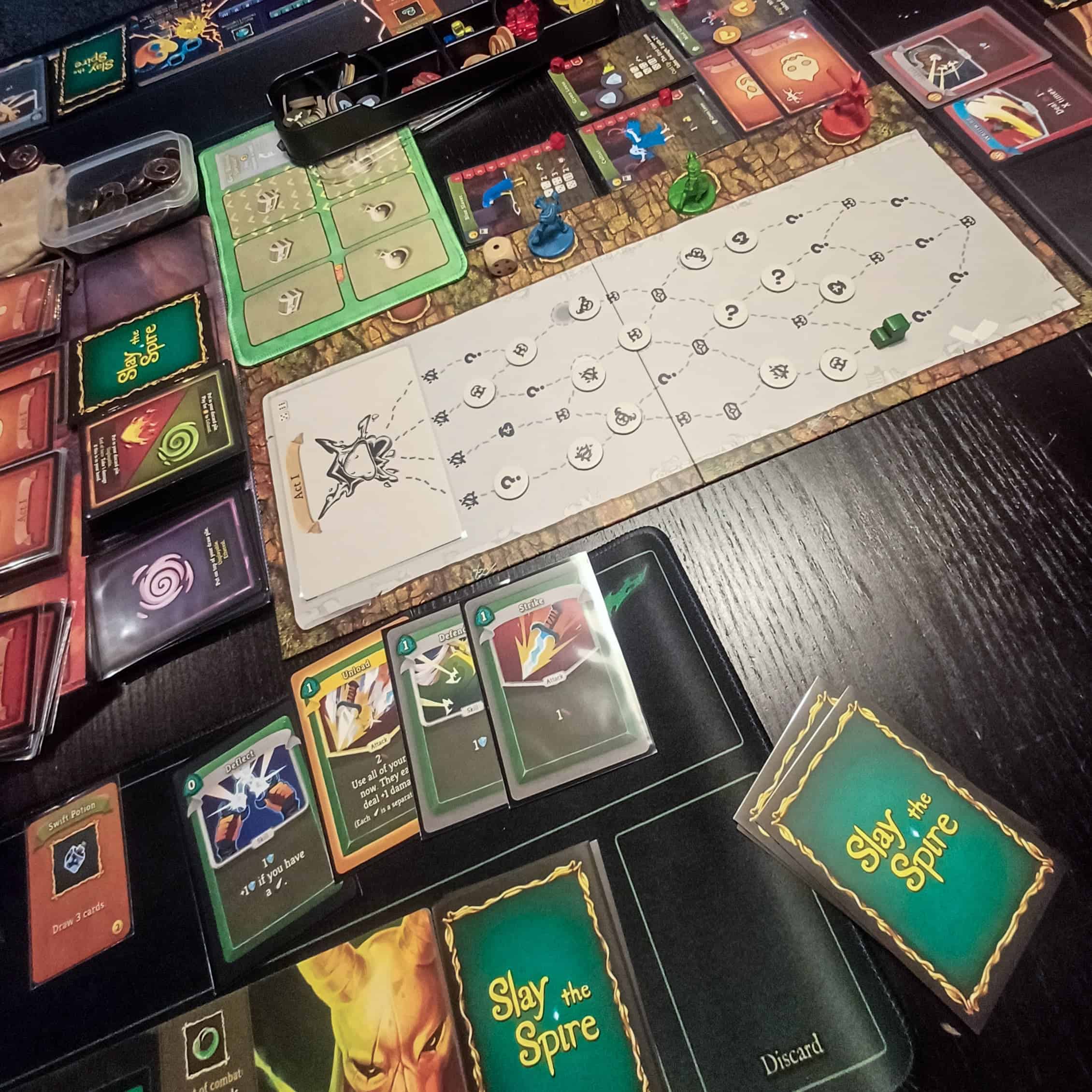 Slay the Spire levällään pöydällä. Etualalla Silent-hahmon kortteja, taustalla paljon erilaisia komponentteja. Oikealla näkyy vähän Ironcladin kortteja.