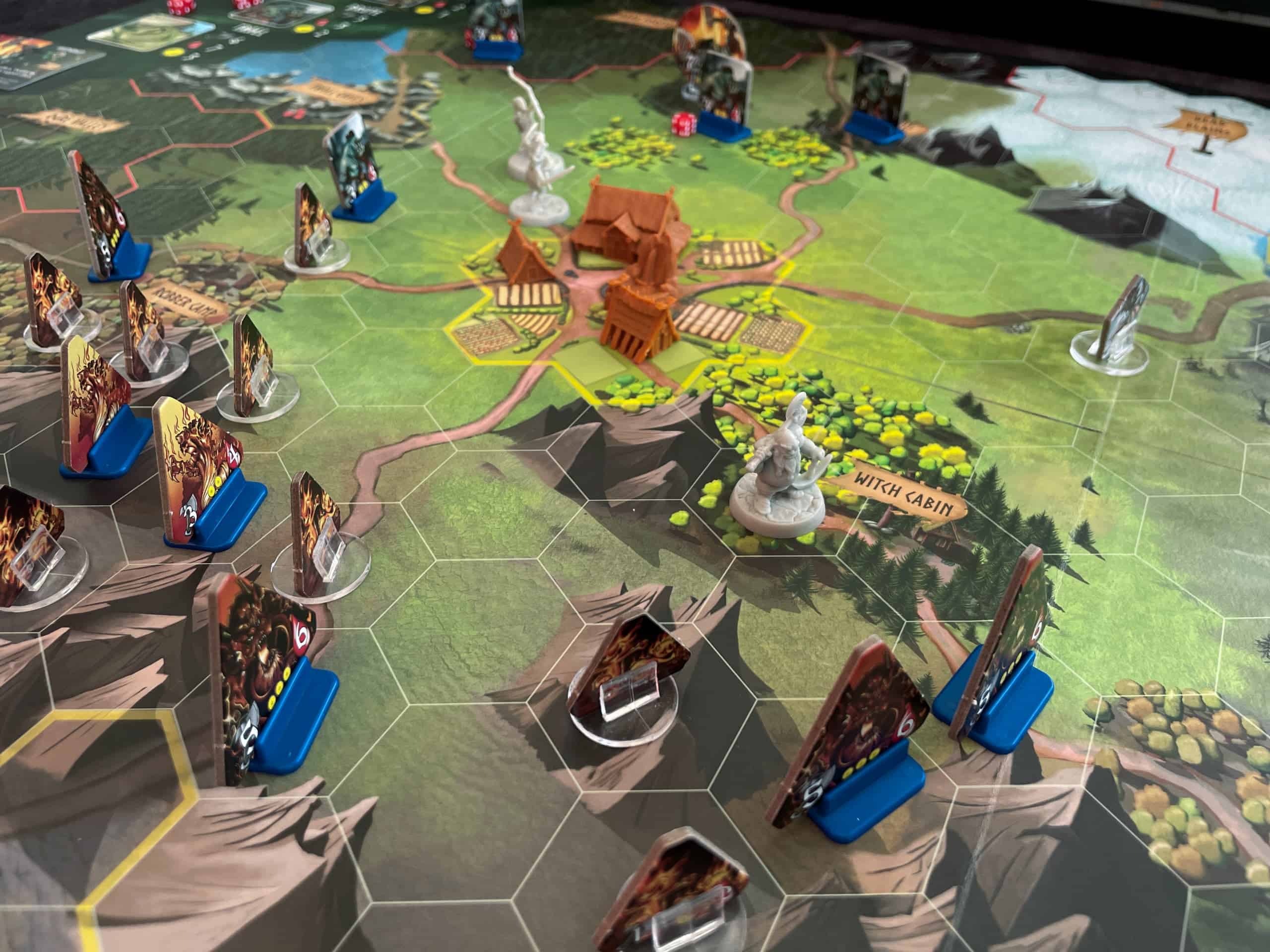 Lähikuva pelilaudasta. Keskellä on kylä, jossa on neljä rakennusta, joka puolelta lähestyy hirviöitä, ja etualalla sankareista kääpiö on torjumassa hyökkäystä  kahden muun sankarin ollessa kylän toisella puolella.