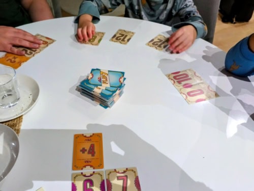 Flip 7 -peli menossa, pelaajia pöydän äärellä kortteineen. Pelissä näkyy olevan mukana neljä pelaajaa, joista kaksi on pieniä lapsia. Pakka kortteja on pöydän keskellä.