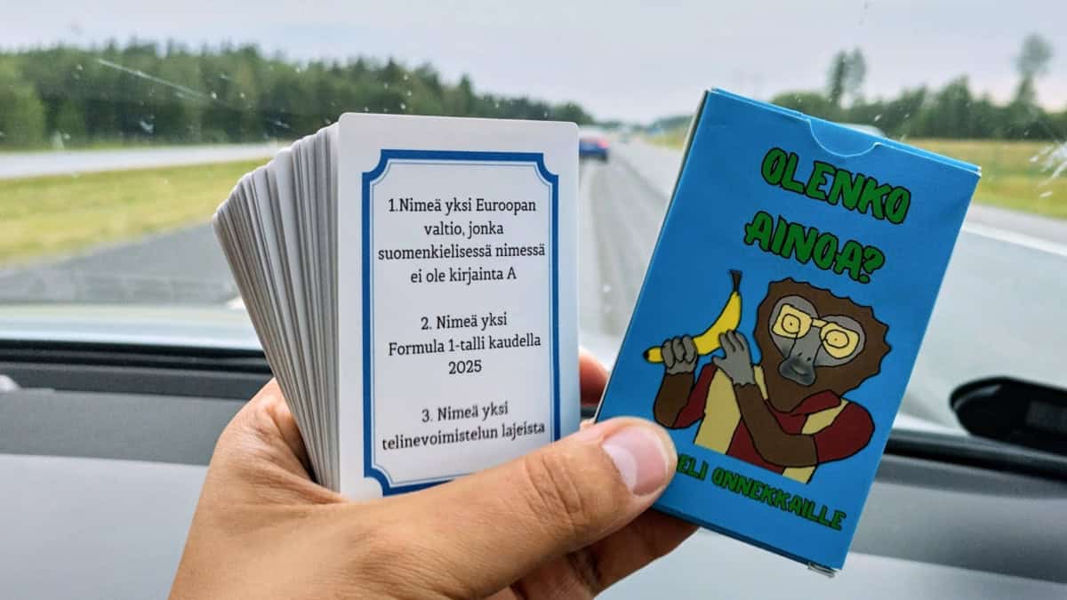 Olenko ainoa? -pelin korttipakka ja pahvinen kotelo kädessä. Kuva on otettu autossa, taustalla näkyy moottoritietä auton tuulilasin läpi. Kotelo on sininen ja siinä on piirros banaania pitelevästä paviaanista. Korttipakan päällimmäisessä kortissa näkyy kolme kysymystä.