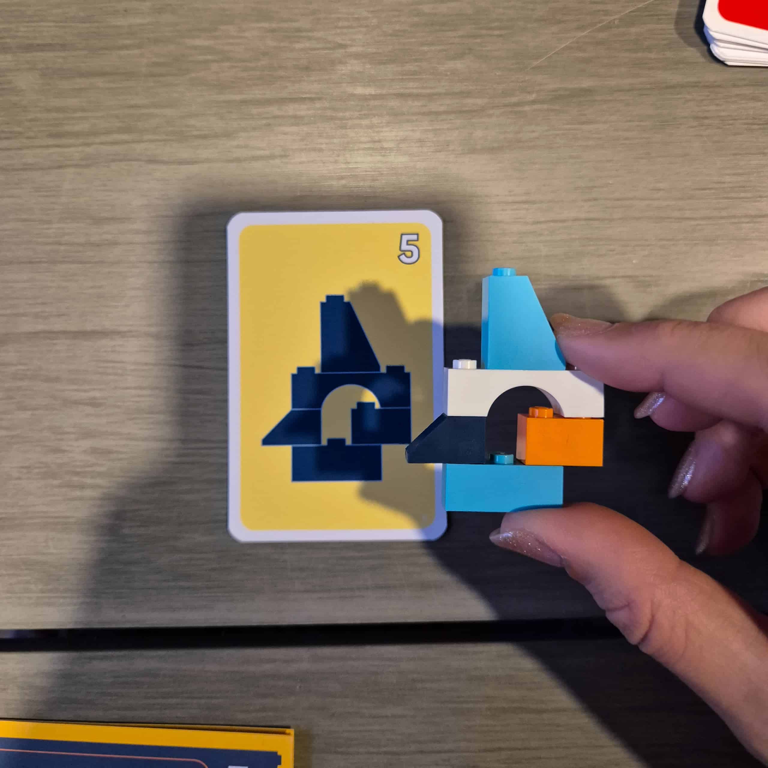 Taustalla tehtäväkortti, jossa on palikkarakennelman muoto, päällä kädessä pideltynä valmis rakennelma.