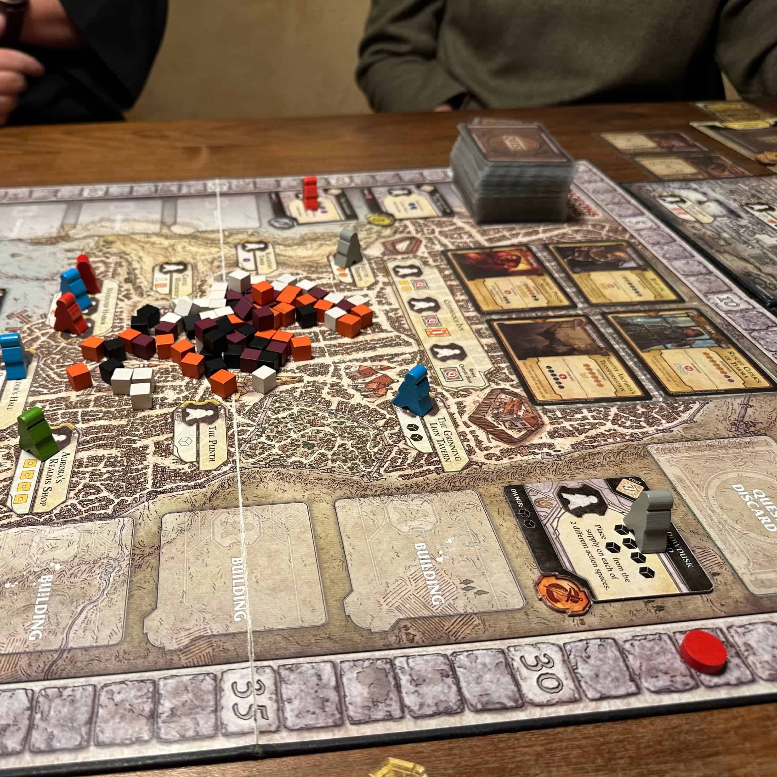 Lähikuva Lords of Waterdeepin pelaajalaudasta, jossa keskellä on läjä puukuutioita, vasemmalla toimintaruutuja, joissa on pelaajien nappuloita ja oikealla kerättävät questit.