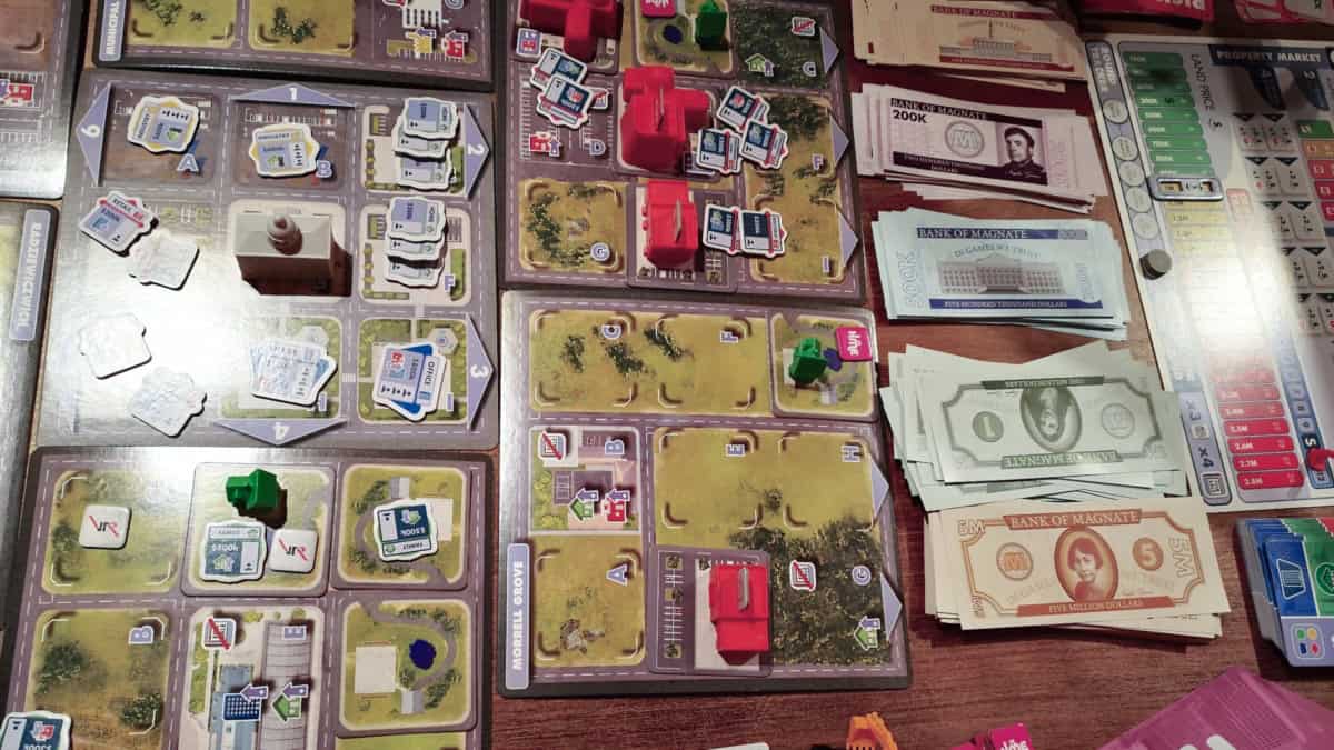 Magnate: The First Cityn pelilautaa, jossa on rakennuksia ja erilaisia pahvimarkkereita. Laudan vieressä on pankki paperirataa ja erillinen pelilauta, jossa on kiinteistömarkkinoiden tilaa seuraavia ratoja.