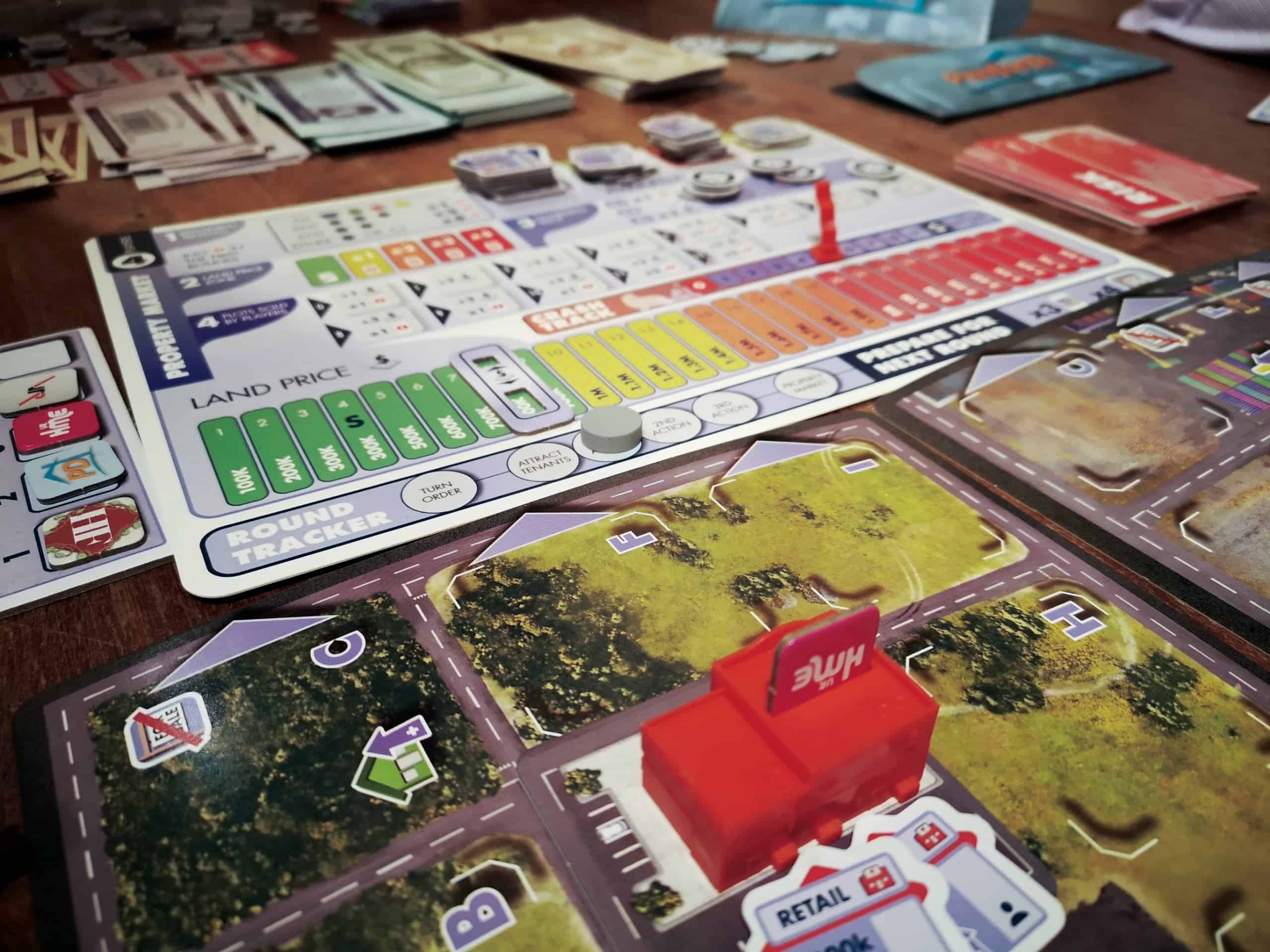 Magnaten kiinteistömarkkinalauta, jossa maan hinta -mittari on vielä alhaisella tasolla, mutta pörssiromahdusmarkkeri on jo yli puolen välin. Etualalla näkyy pelilautaa, jossa on punainen liikerakennus.