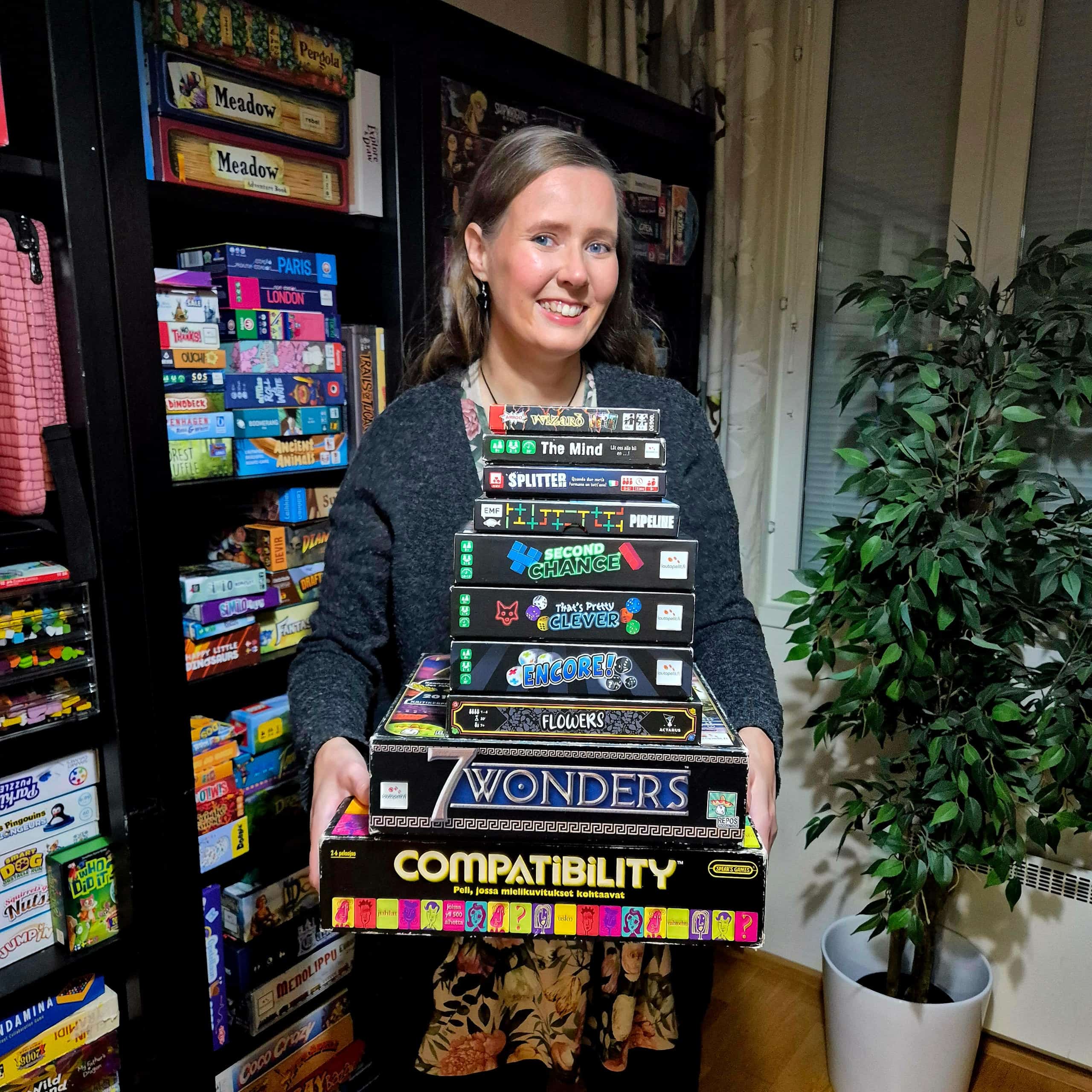 Vilma peleillä täytetyn kirjahyllyn edessä, sylissään nippu mustia pelejä: Wizard, The Mind, Splitter, Pipeline, Second Chance, That's Pretty Clever, Encore!, Flowers, 7 Wonders ja Compatibility.