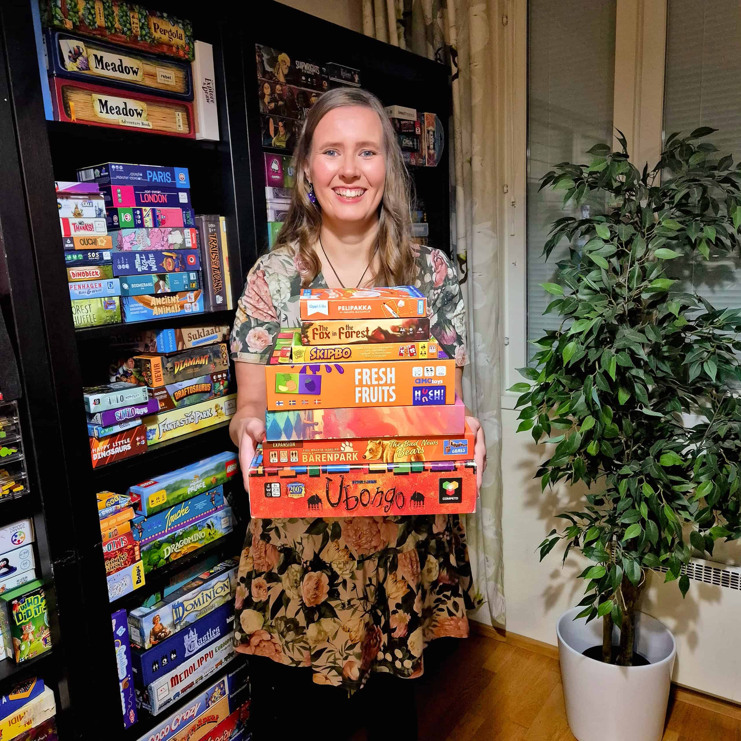 Vilma pelihyllyn ja limoviikunan edessä, sylissään pino oransseja pelejä: Pelipakka, The Fox in the Forest, Skip-Bo, Fresh Fruits, Canvas, Bärenparkin lisäosa ja Ubongo.