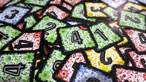 Flowersin neliönmuotoisia kukkakuvioisia numerokortteja sekalaisena läjänä pöydällä.