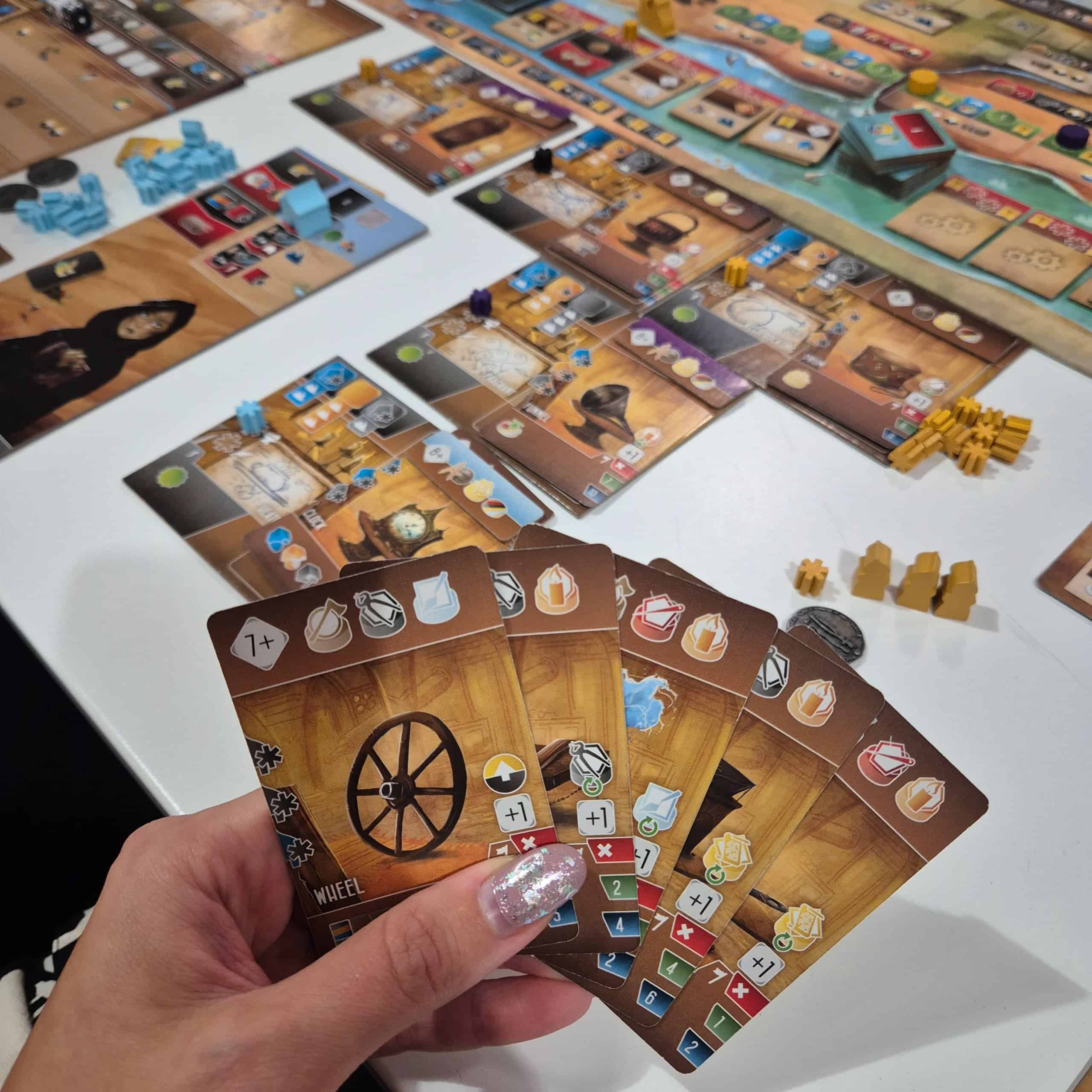 Lähikuva kädestä pitelemässä Inventors of the South Tigrisin kortteja. Taustalla näkyy pelilautaa ja pelaajan komponentteja.