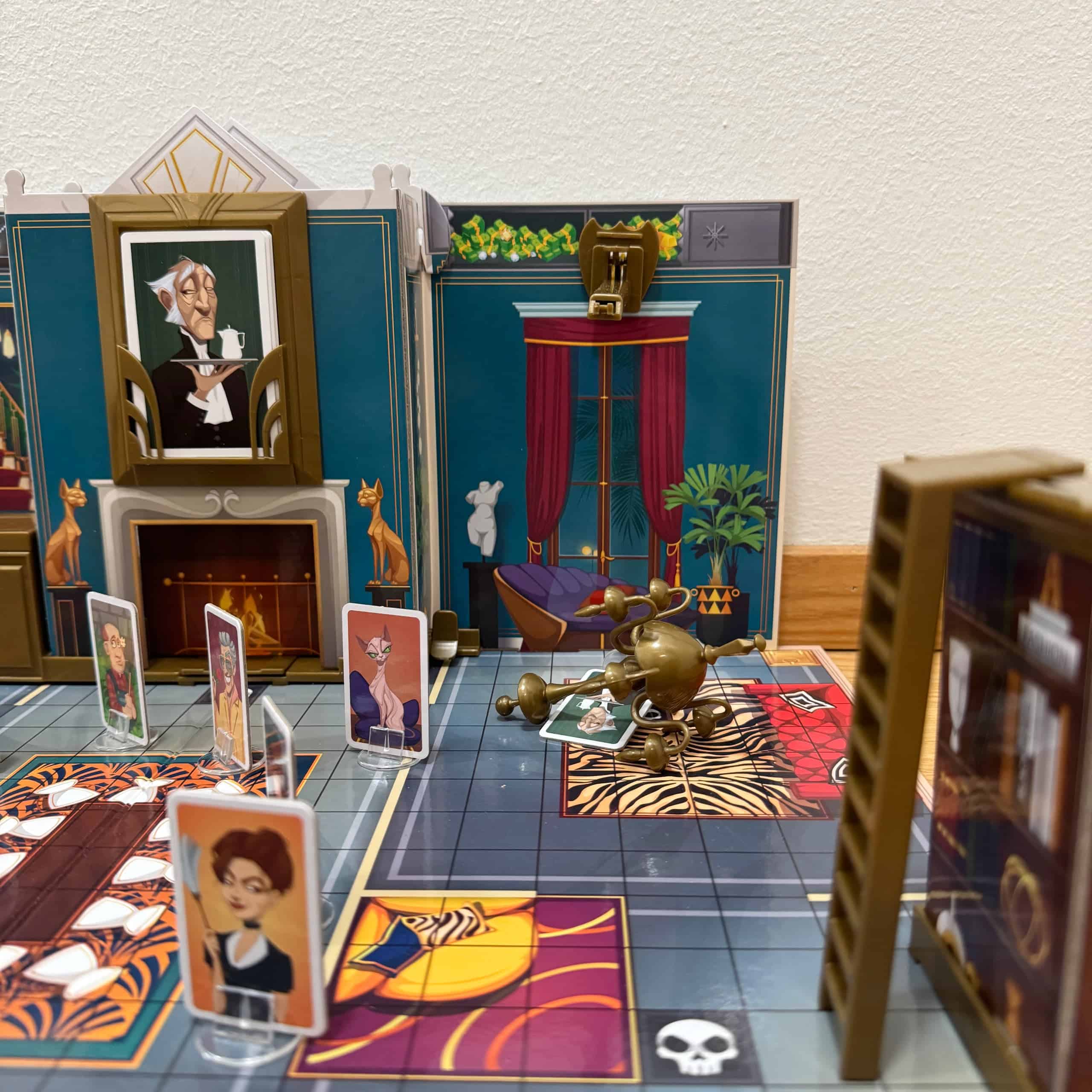 Lähikuva pelin 3d-pelilaudasta, jossa hovimestarinappula on jäänyt pudonneen kristallikruunun alle. Kissanappula katselee vierestä.