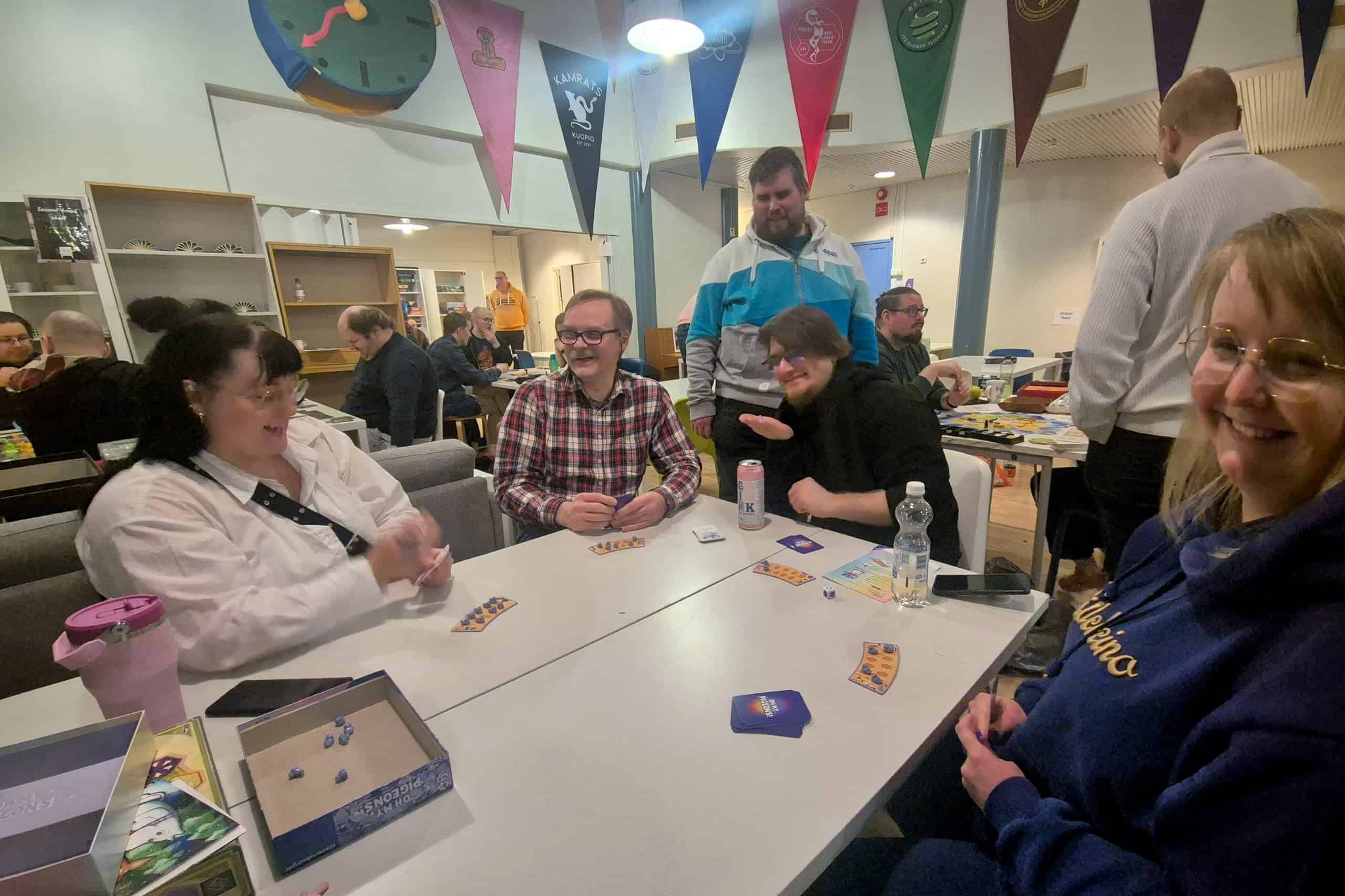 Neljä ihmistä pöydän äärellä, viides seisoo vieressä katsomassa. Pöydällä Oh My Pigeons! -peli.