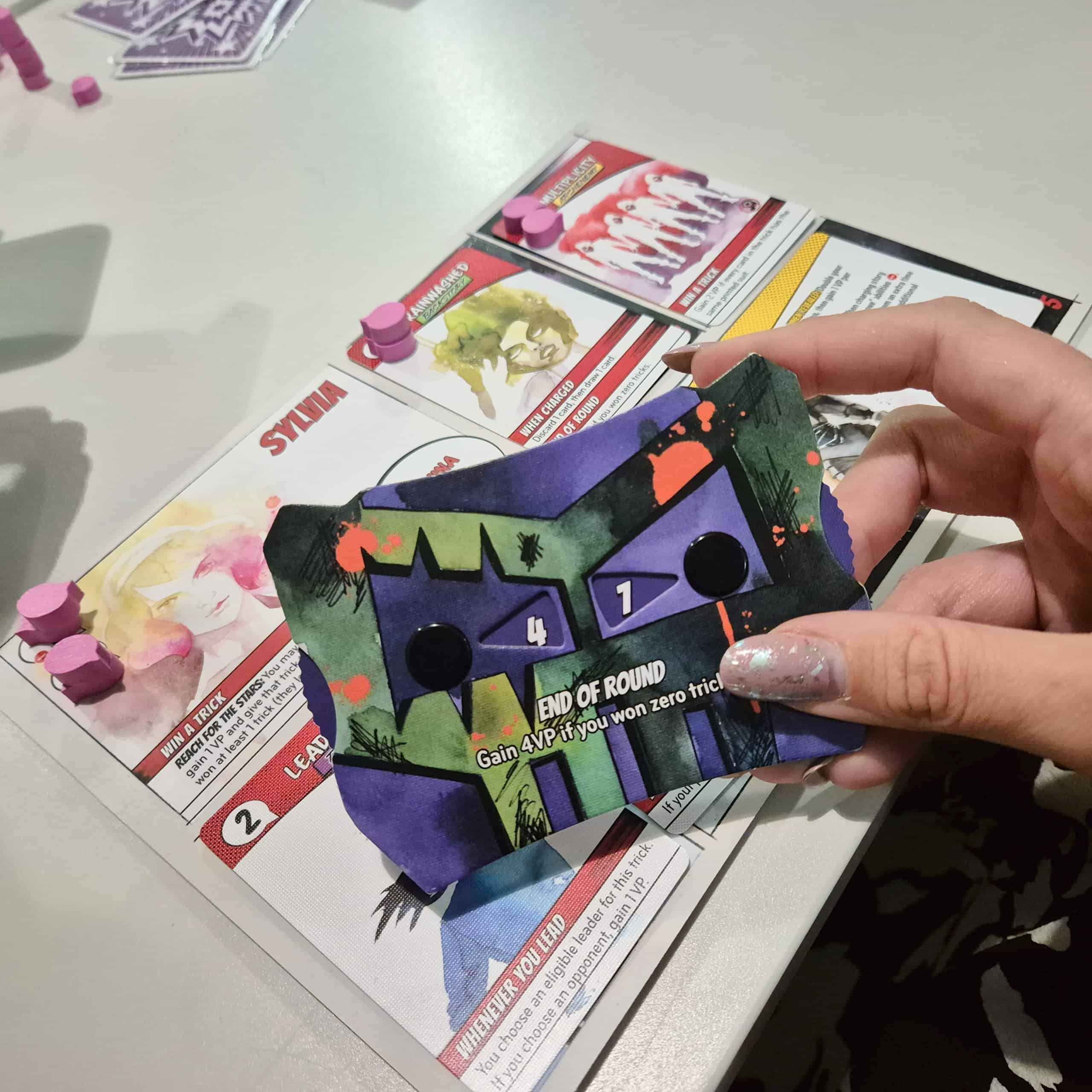 Lähikuva kädestä pitelemässä Origin Storyn pistelaskuria, taustalla Sylvia-hahmon pelilauta.