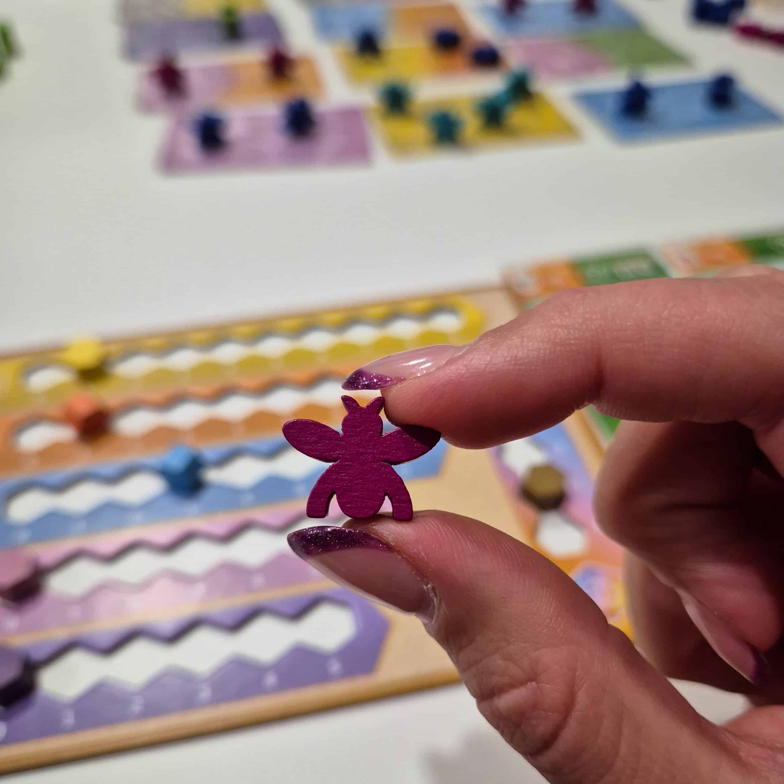 Sormet pitelemässä violettia mehiläisnappulaa. Taustalla näkyy sumeana pelilautoja.