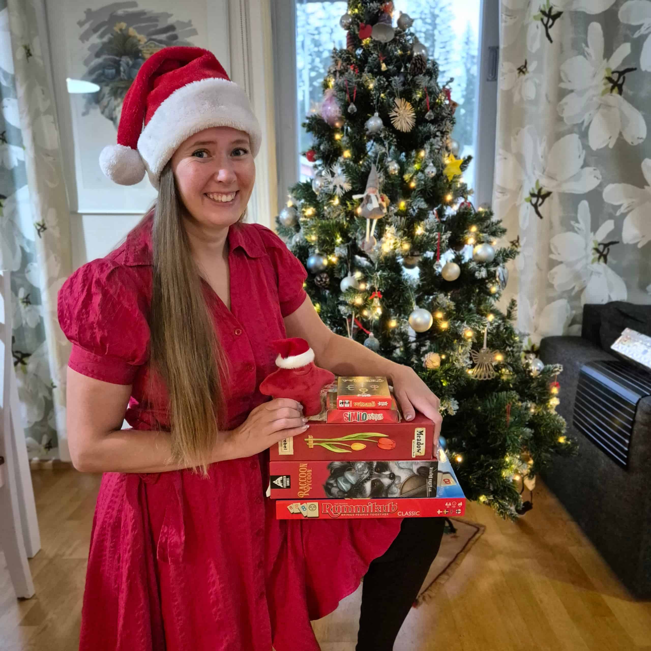 Vilma tonttuhatussa ja punaisessa mekossa, taustallaan joulukuusi. Vilmalla on sylissä punaisia pelilaatikoita: Wizard, Similo Animals, Meadow, Raccoon Tycoon ja Rummikub.