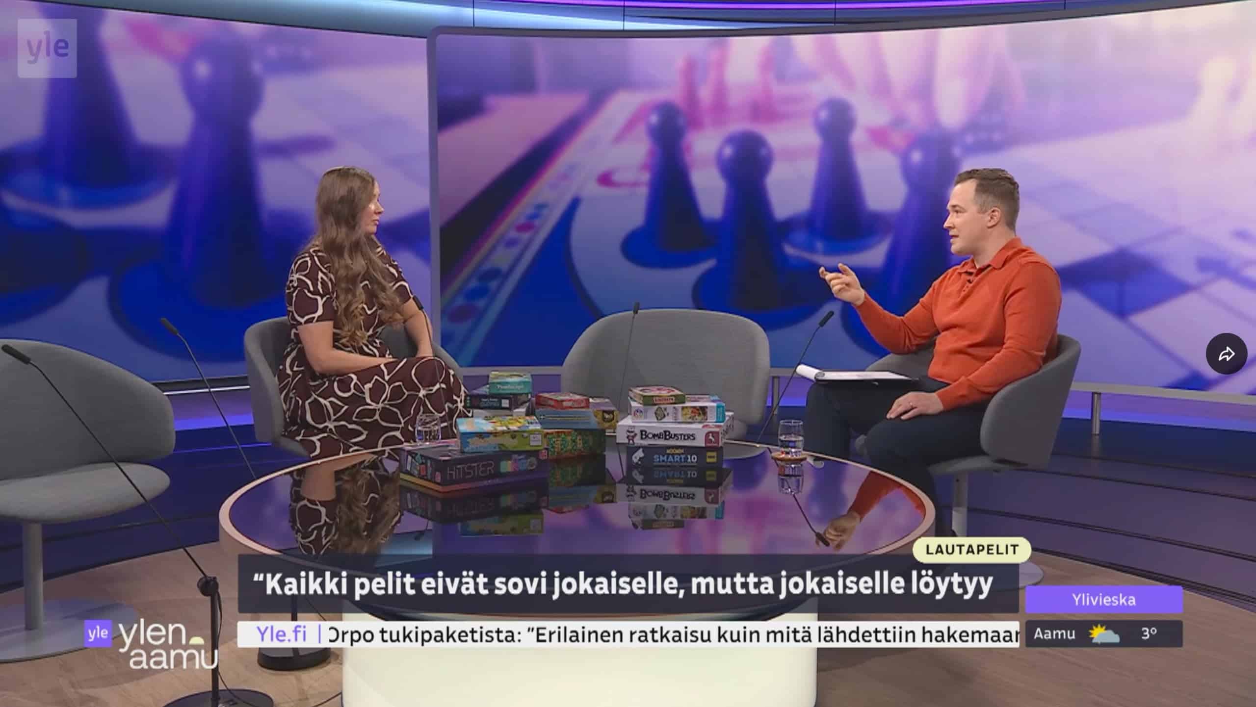 Vilma vierailemassa Ylen Aamussa. Studiossa Vilma ja haastattelija, välissään pöydällä paljon lautpaelejä. Taustalla on kuvia pelinappuloista.