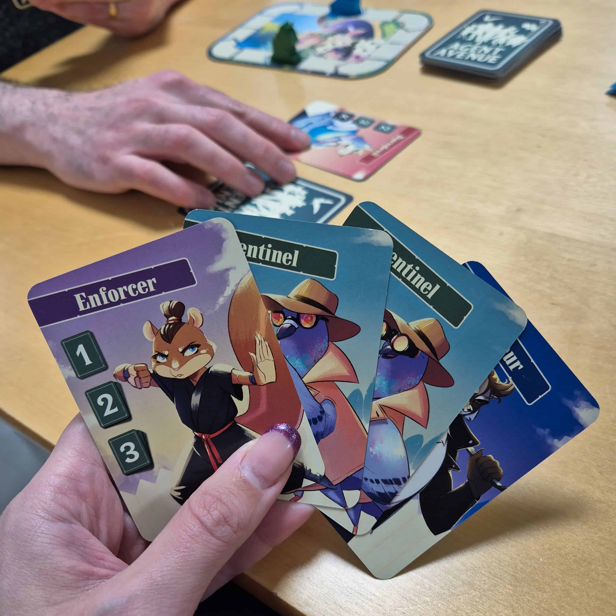 Käsi pitelemässä neljää Agent Avenuen korttia, joissa on kuvattuna ihmismäisiä eläinhahmoja. Taustalla näkyy toisen pelaajan käsi ja kaksi korttia, toinen kuvapuoli ylöspäin ja toinen kuvapuoli alaspäin.