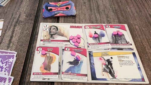 Origin Storyn hahmo Nnamdi. Hahmolaudalla on neljä korttia: Backstory-kortti "Fateful choice", voimakortti "Invisibility", liittolainen "Serpio" ja kyky "Leadership". Supersankarikorttina on "Stygia".