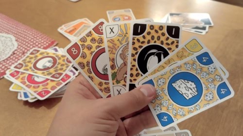 Pelaajan kädessä erilaisia pitsatäytekortteja: sieni, fantasia, oliivi, ananas ja homejuusto. Pöydällä käden takana näkyy korttipinoja.