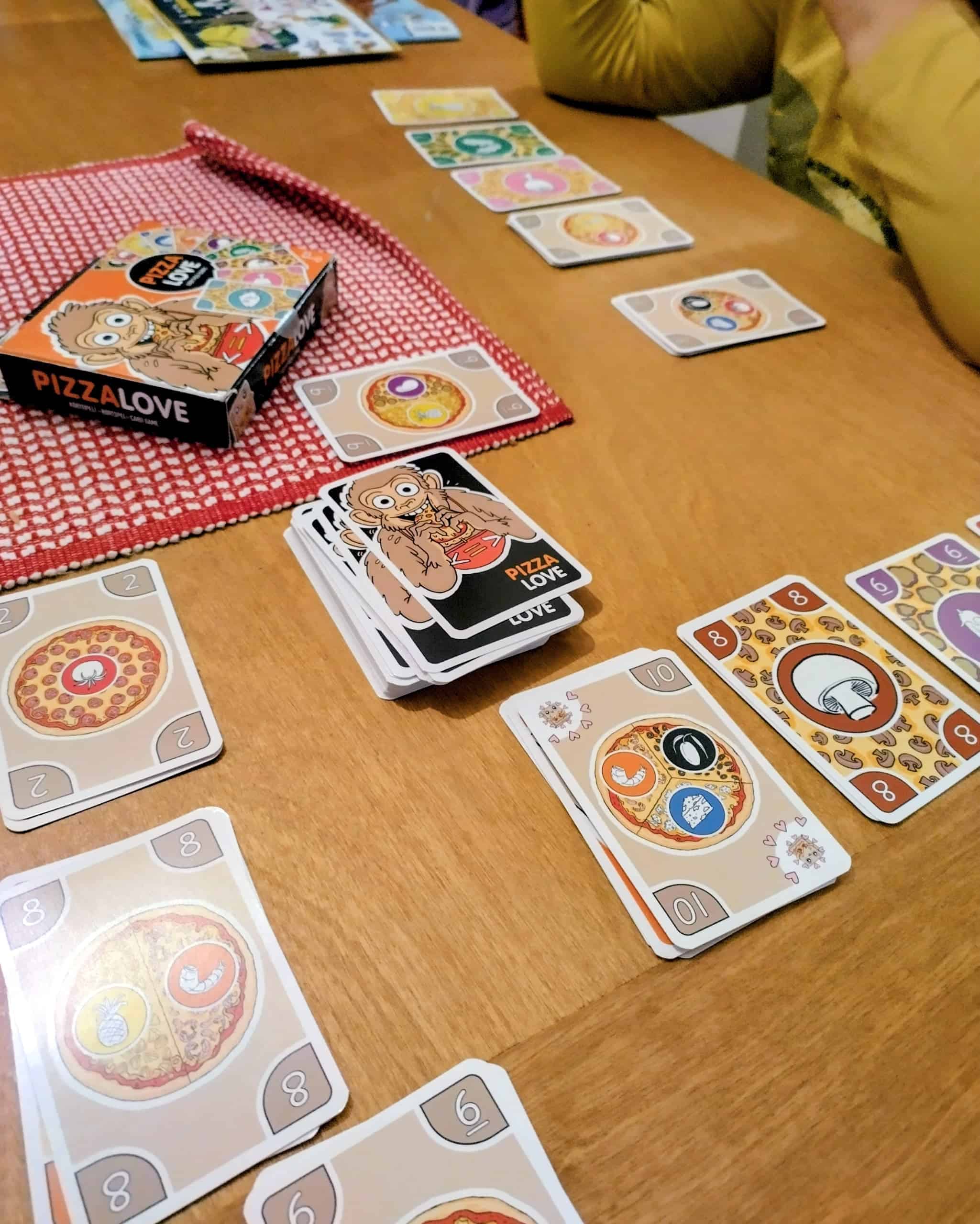 Peli menossa. Pelaajien edessä on rivit täytekortteja varastossa ja pino pizzakortteja.