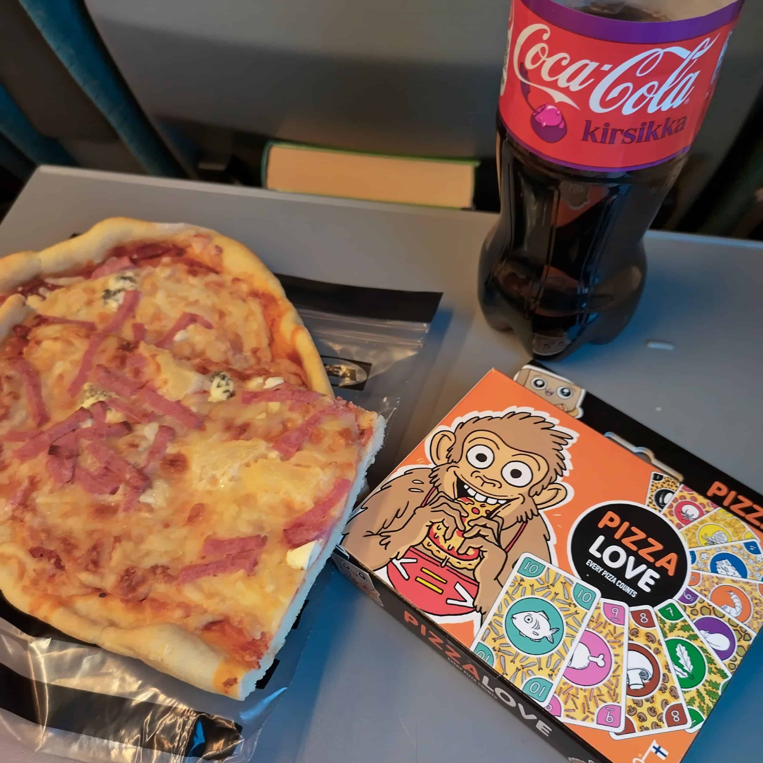 Junan pöydällä pala kinkku-aurajuustopizzaa, Pizza Loven paketti ja 1,5 litran pullo kirsikkakokista.