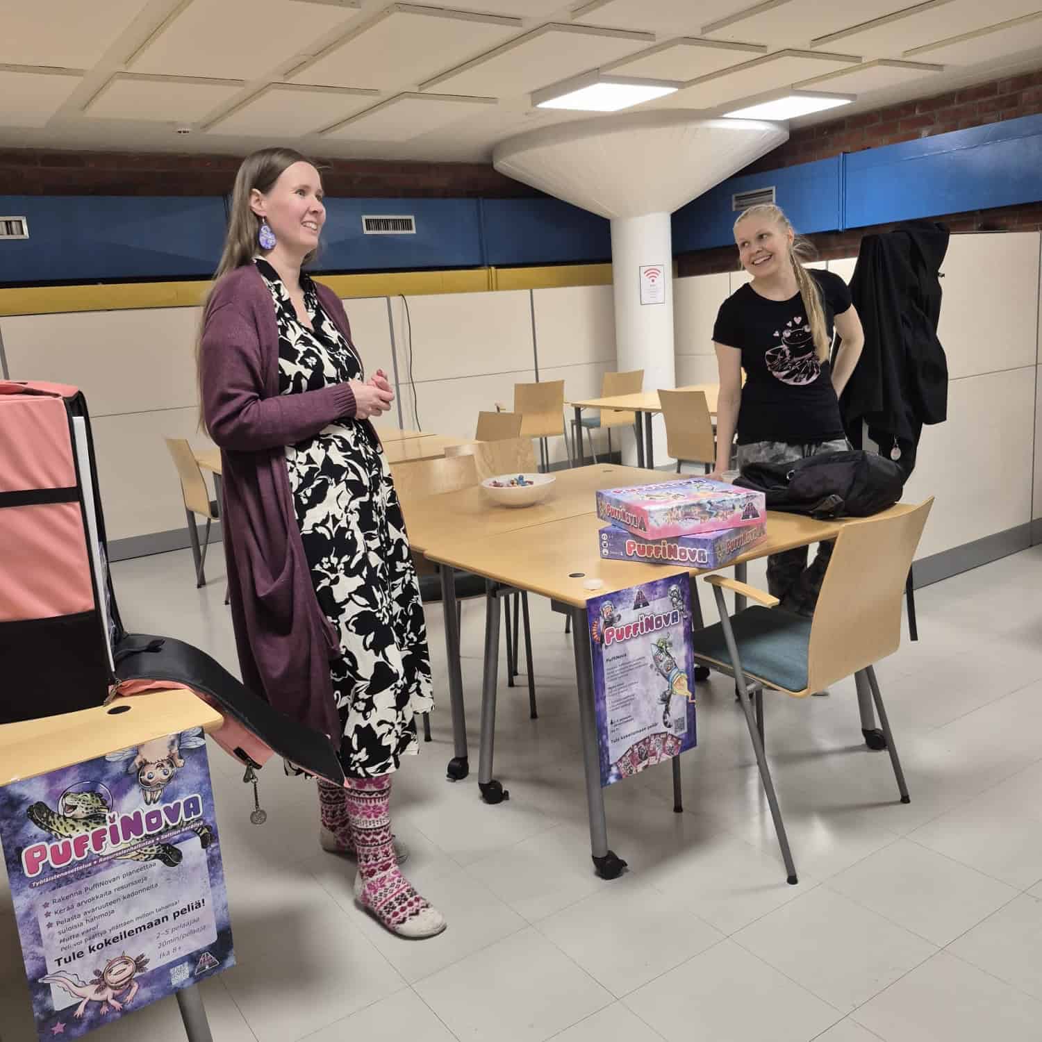Vilma ja Jasu seisomassa pöydän molemmin puolin. Pöydällä on kaksi PuffiNova-pelilaatikkoa. Pöydässä ja viereissä pöydässä on kiinni PuffiNovan pelidemoa mainostavat julisteet.