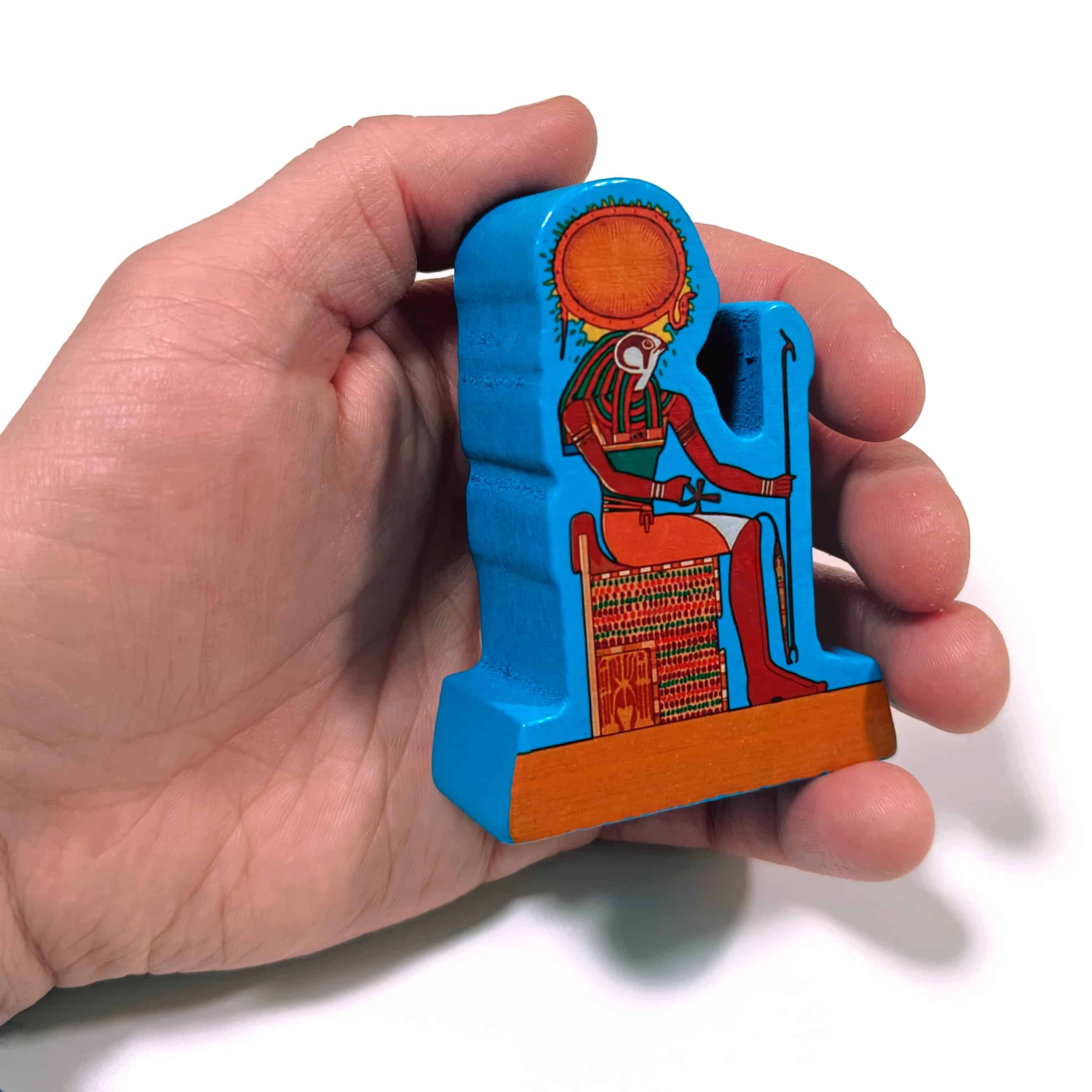 Käsi pitelemässä pelin puista Ra-patsasta, joka kuvaa Ra-jumalaa istumassa valtaistuimellaan. Patsas on aikuisen miehen käteen sopivan kokoinen.