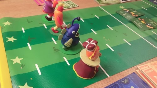 Pelin neljä maskottihahmoa rivissä valmiina kilpajuoksuun vihreää juoksurataa kuvaavalla pelilaudalla.