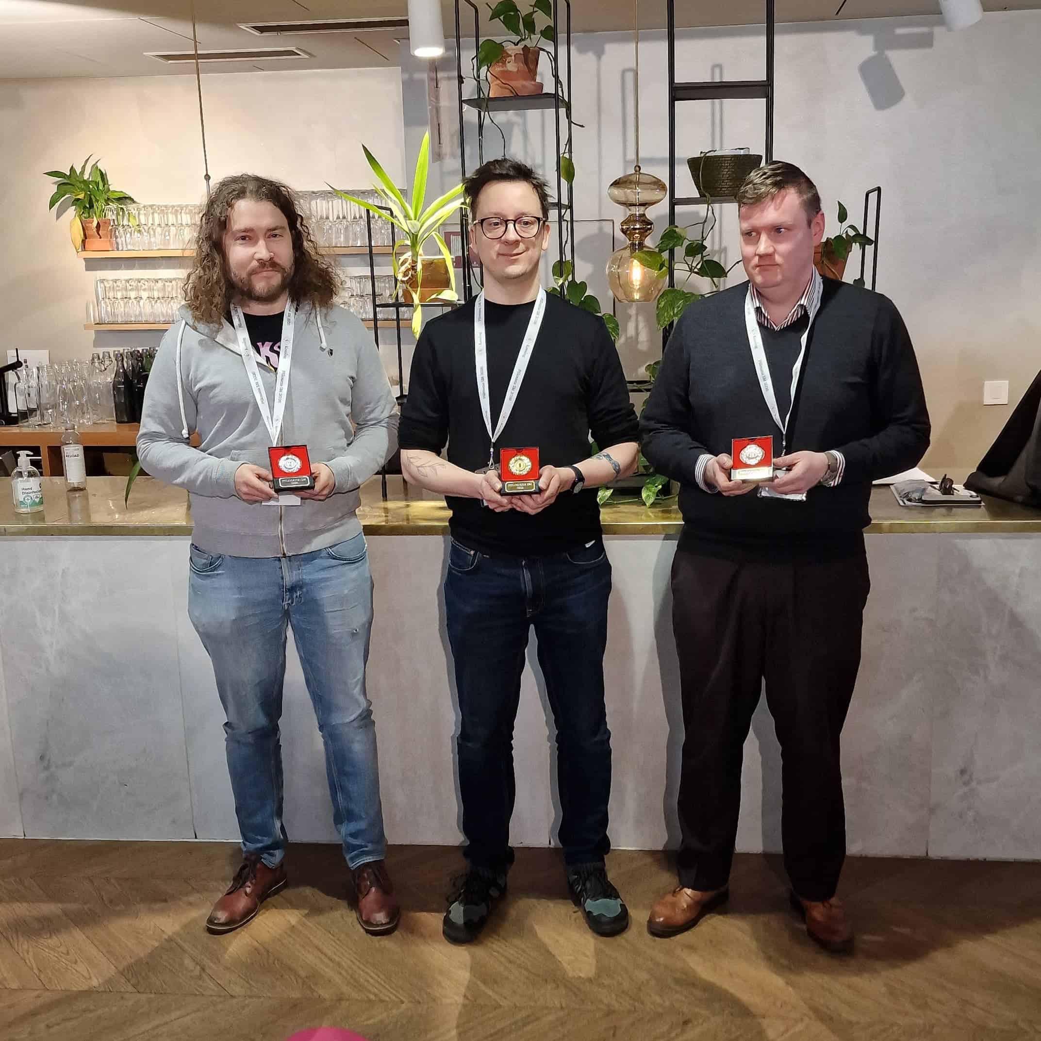 Kolme vierekkäin seisovaa miestä ravintolan tiskin edessä. Kaikilla on kädessään rasiassa oleva mitali.