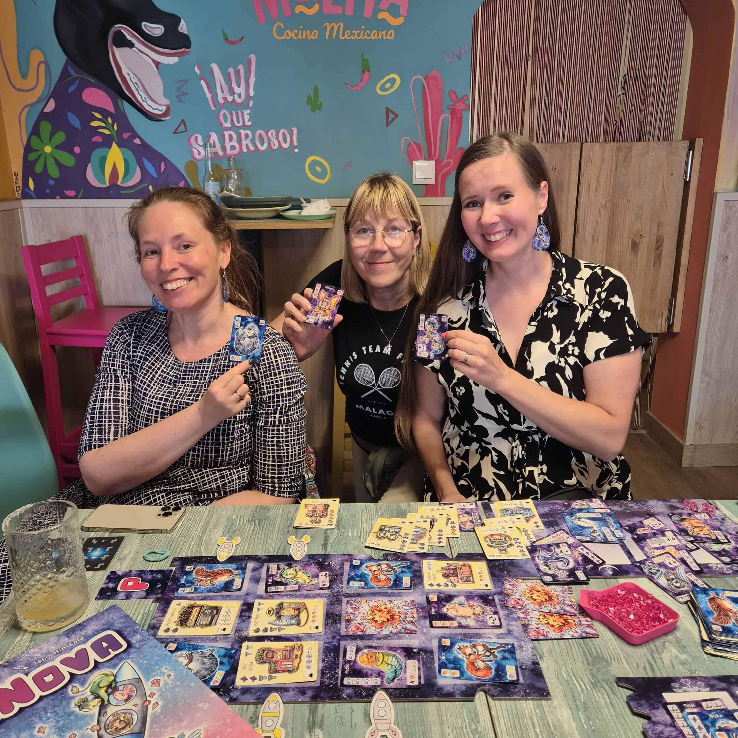 Kolme naista ravintolassa pöydän ääressä. Kaikilla on kädessään PuffiNova-kortti. Pöydällä naisten edessä on levittettynä PuffiNova-peli.