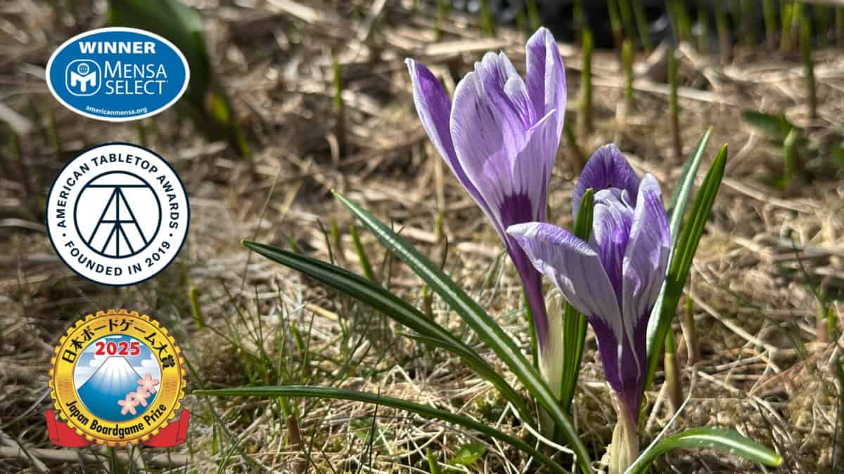 Violetti- ja valkoraidallisia krookuksia maassa. Vasemmassa reunassa pinossa Mensa Selectin, ATTA:n ja Japan Board Game Awardin palkintologot.