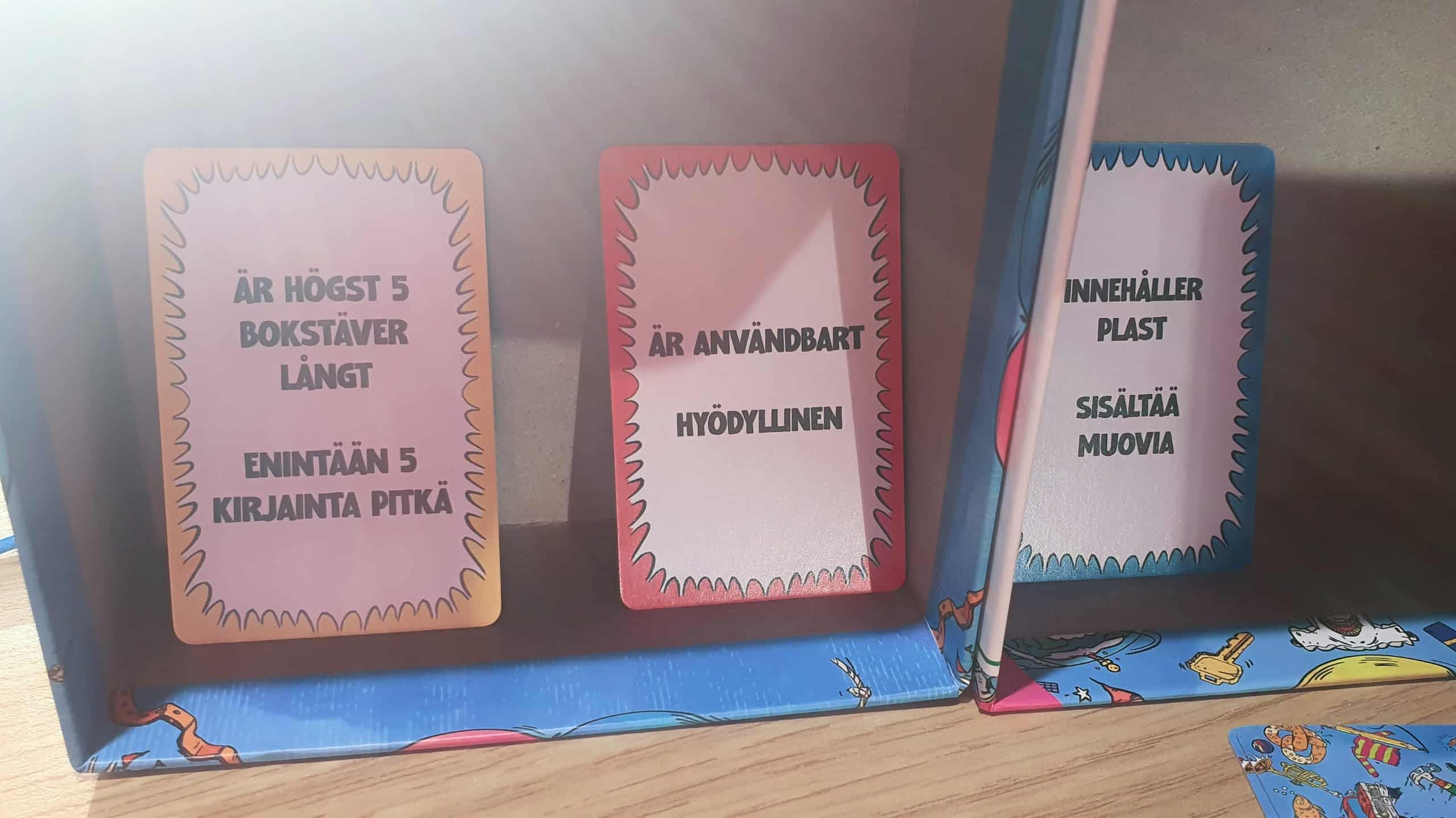 Kolme kategoriakorttia piilossa pelin laatikoiden takana: keltainen "enintään 5 kirjainta pitkä", punainen "hyödyllinen" ja sininen "sisältää muovia".