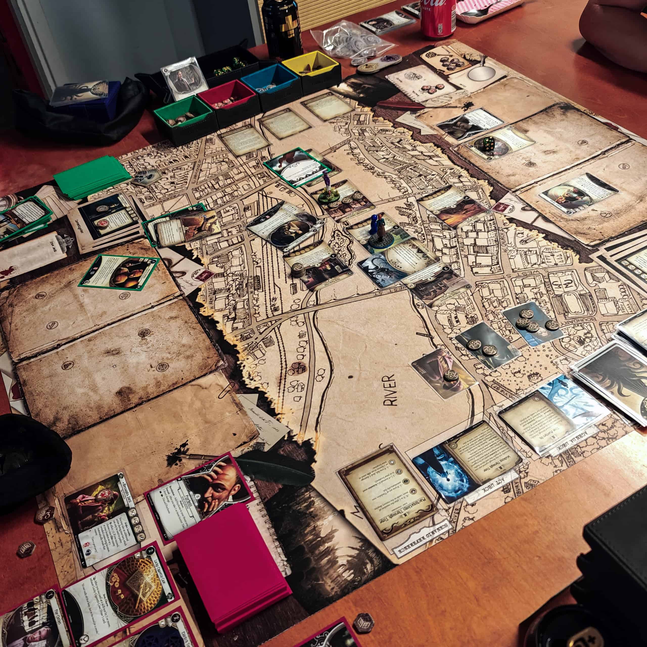 Pöydällä Arkhamin kaupunkia kuvaava Arkham Horror LCG:n pelimatto, jonka päällä on pelin kortteja ja joillain korteilla noppia ja miniatyyrejä.