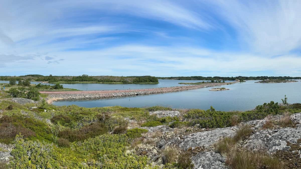 Kesäinen päivä, puolipilvinen taivas Brändön Torsholmaan johtavan tien yllä. Etualalla pientä katajaa kasvavia kallioita.