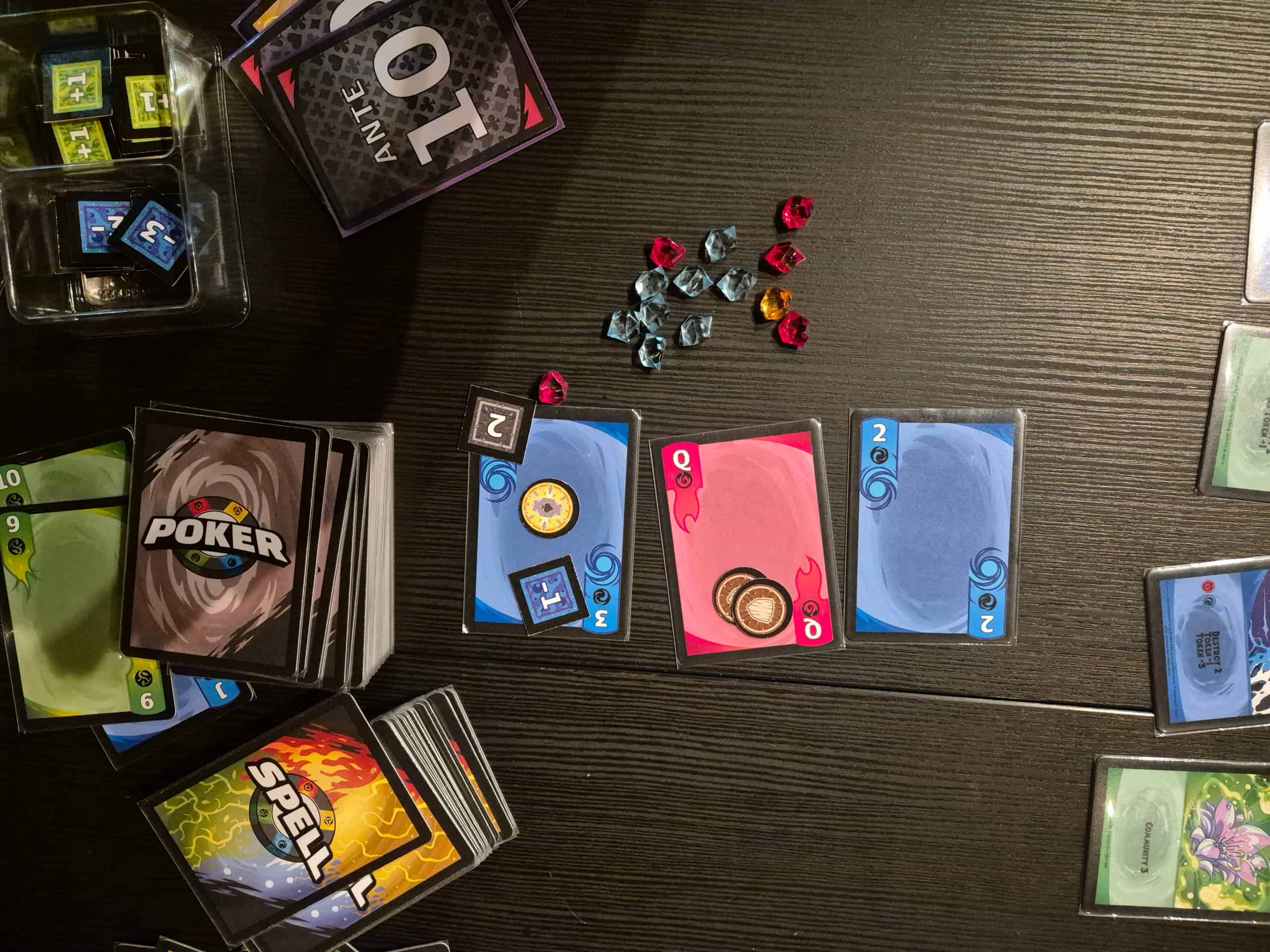 Spell Poker -pelin kortteja pöydällä. Korttien päällä on pahvitokeneita ja vieressä muovijalokiviä.