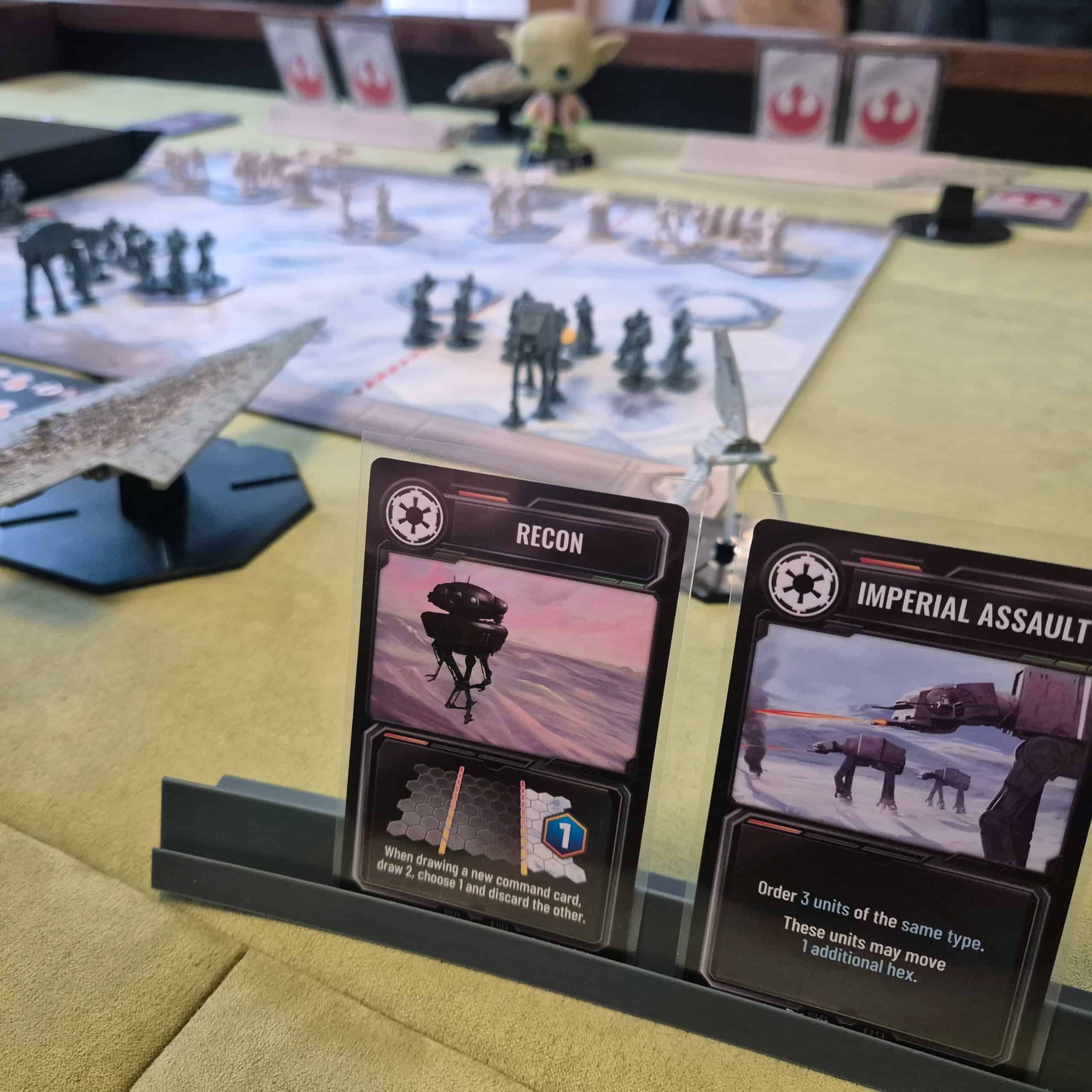 Kaksi Star Wars: Battle of Hothin korttia telineessä etualalla, taustalla pelilauta, jossa sotajoukot taistelevat.