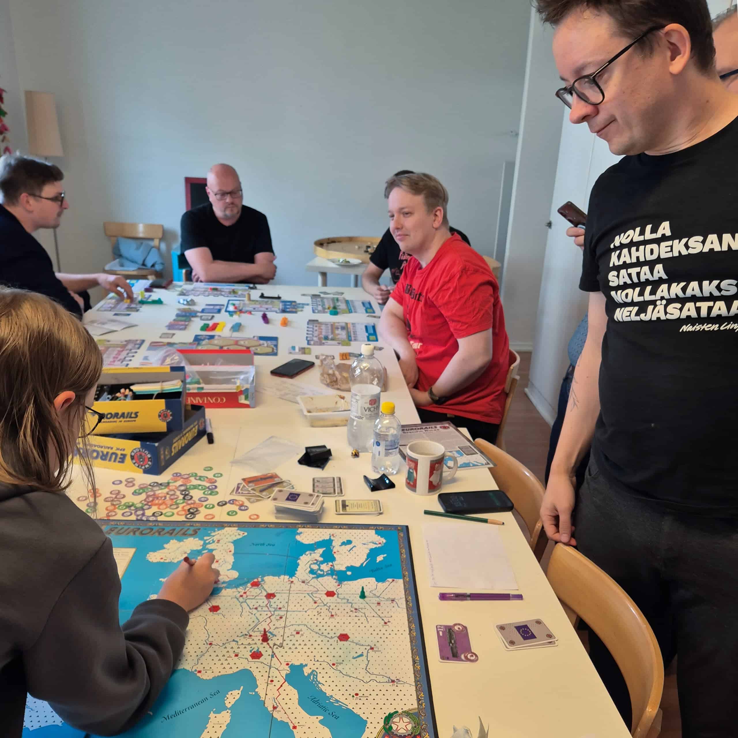 Etualalla Leevi piirtää Eurorails-ratoja ja Mikko seisoo pöydän vieressä vahtimassa, taustalla pelataan Containeria.