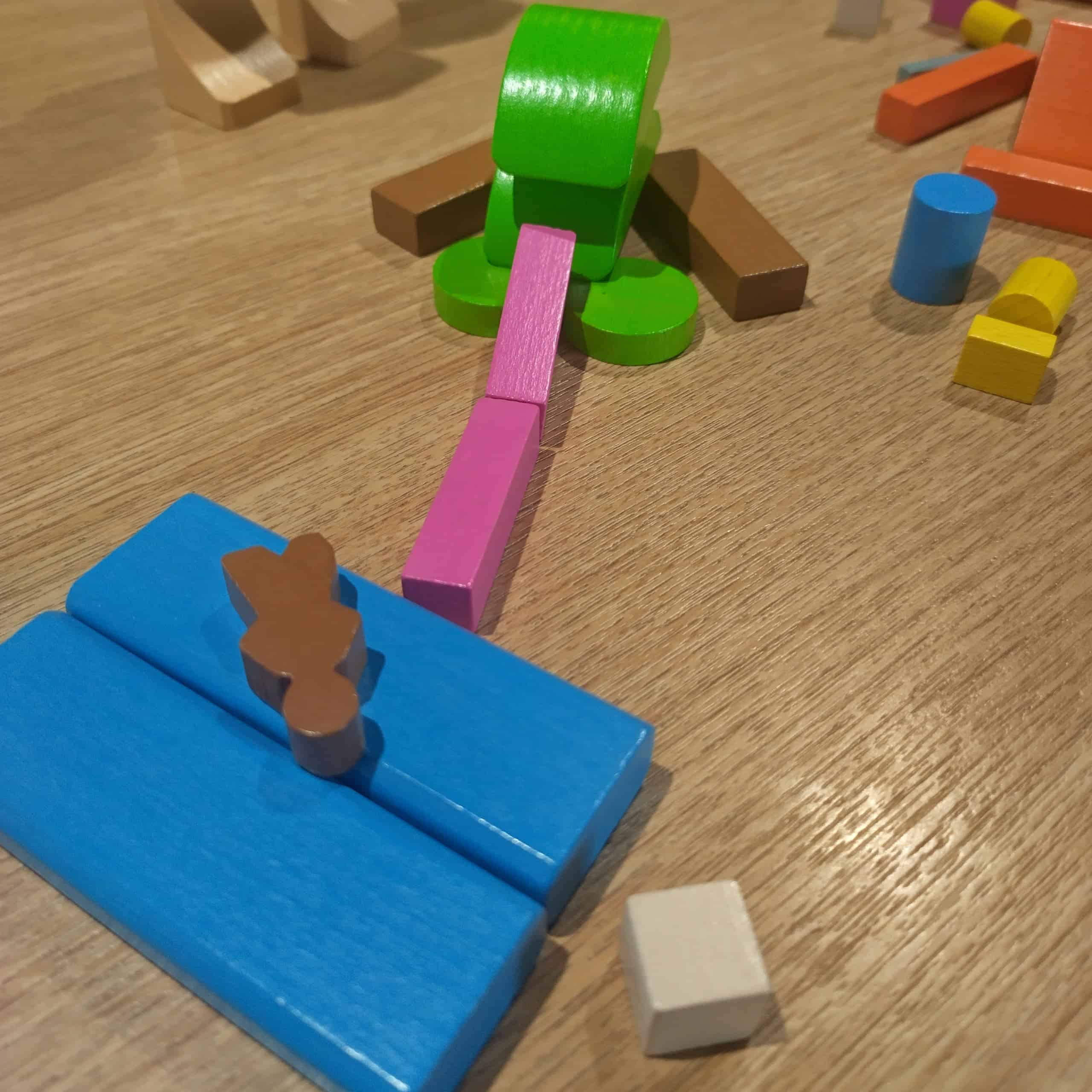 Palikoista rakennettu vihreä sammakko, jolla on pitkä pinkki kieli joka kurottaa kohti sinisten palikoiden päällä olevaan ruskeaa ihmishahmoa.